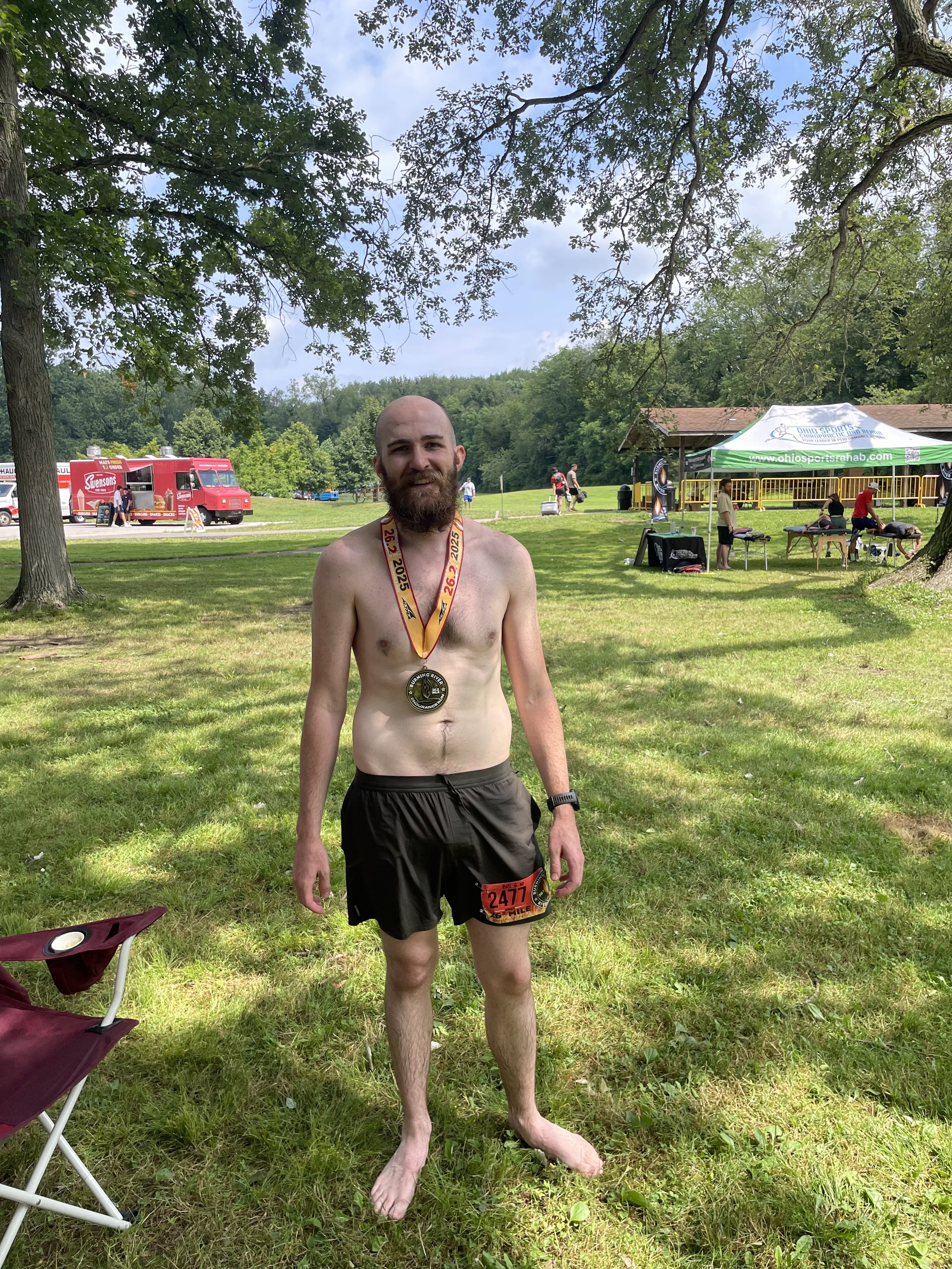 A shirtless man with a medal around his neck and a race bib number 2477 standing on grass in a park after a race, with trees, a food truck, a tent, and people in the background.