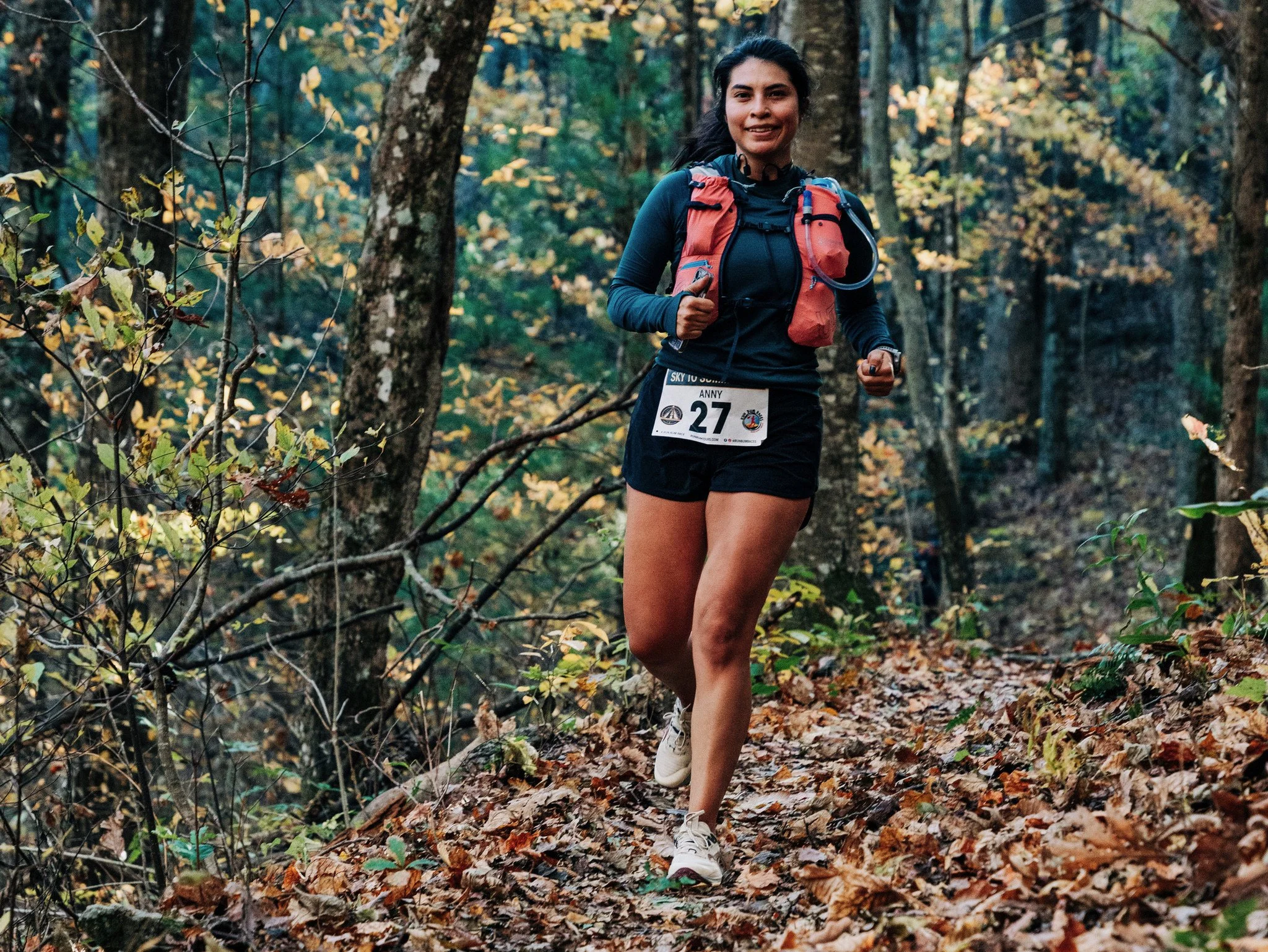 A woman running on a trail in a forest during fall, wearing a black athletic outfit, a red hydration pack, and a race bib number 27.
