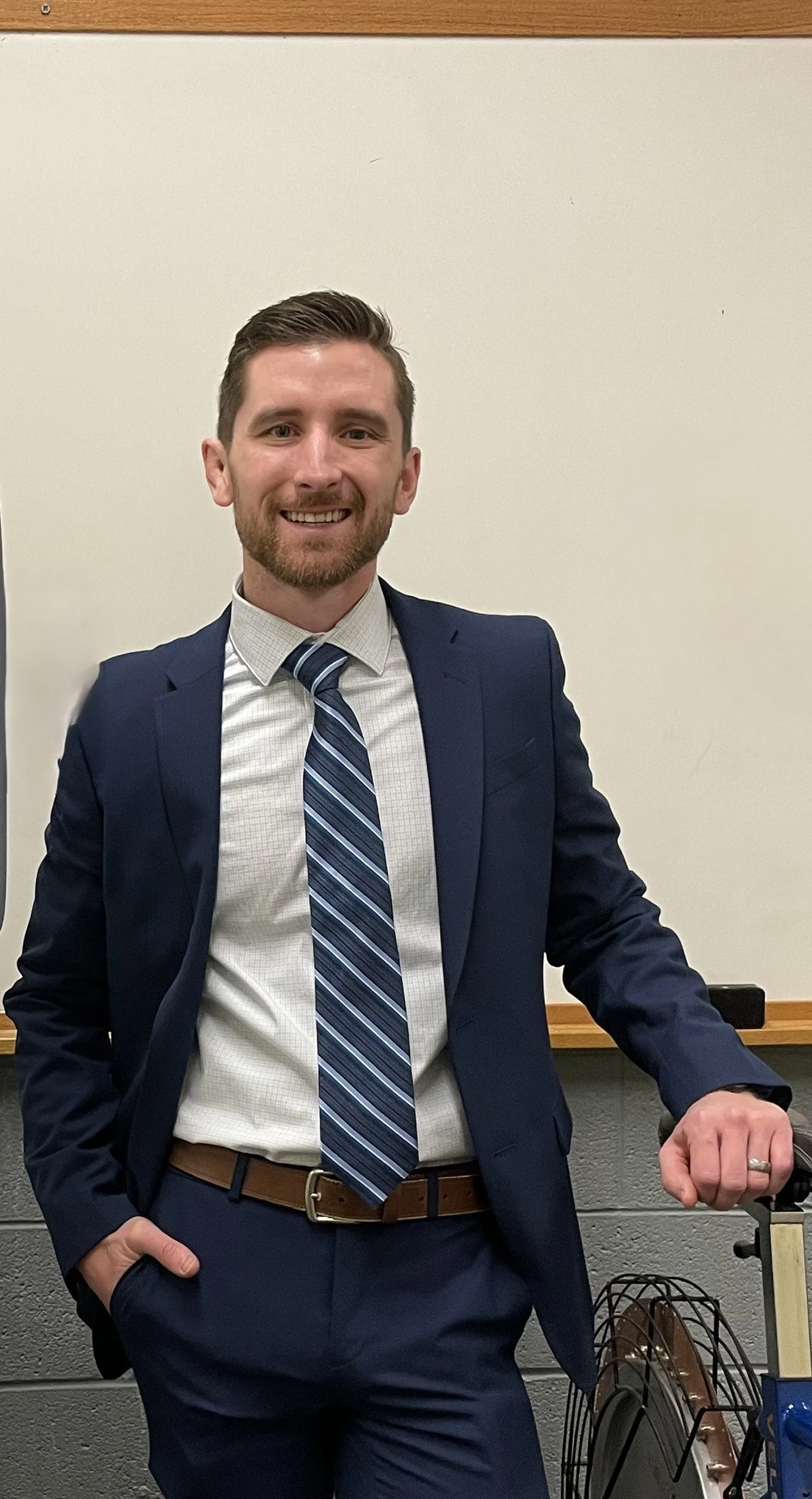 A smiling man in a navy suit, white shirt, and striped blue tie, standing in a classroom or office setting with a whiteboard behind him, one hand in his pocket and the other resting on a piece of equipment.