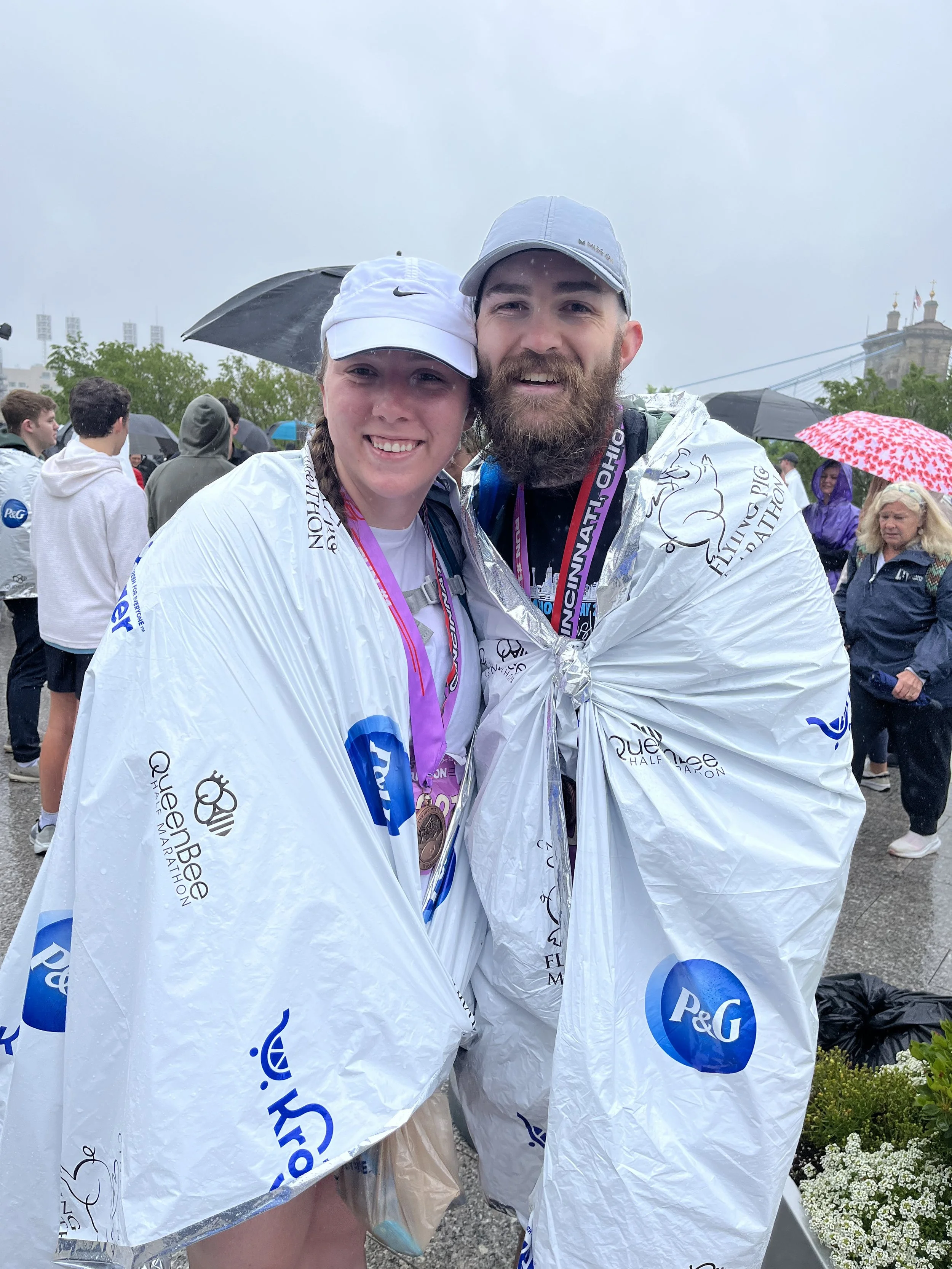 Two runners, a woman and a man, smiling and wearing ponchos after completing a race, with other runners and umbrellas in the background on a rainy day.