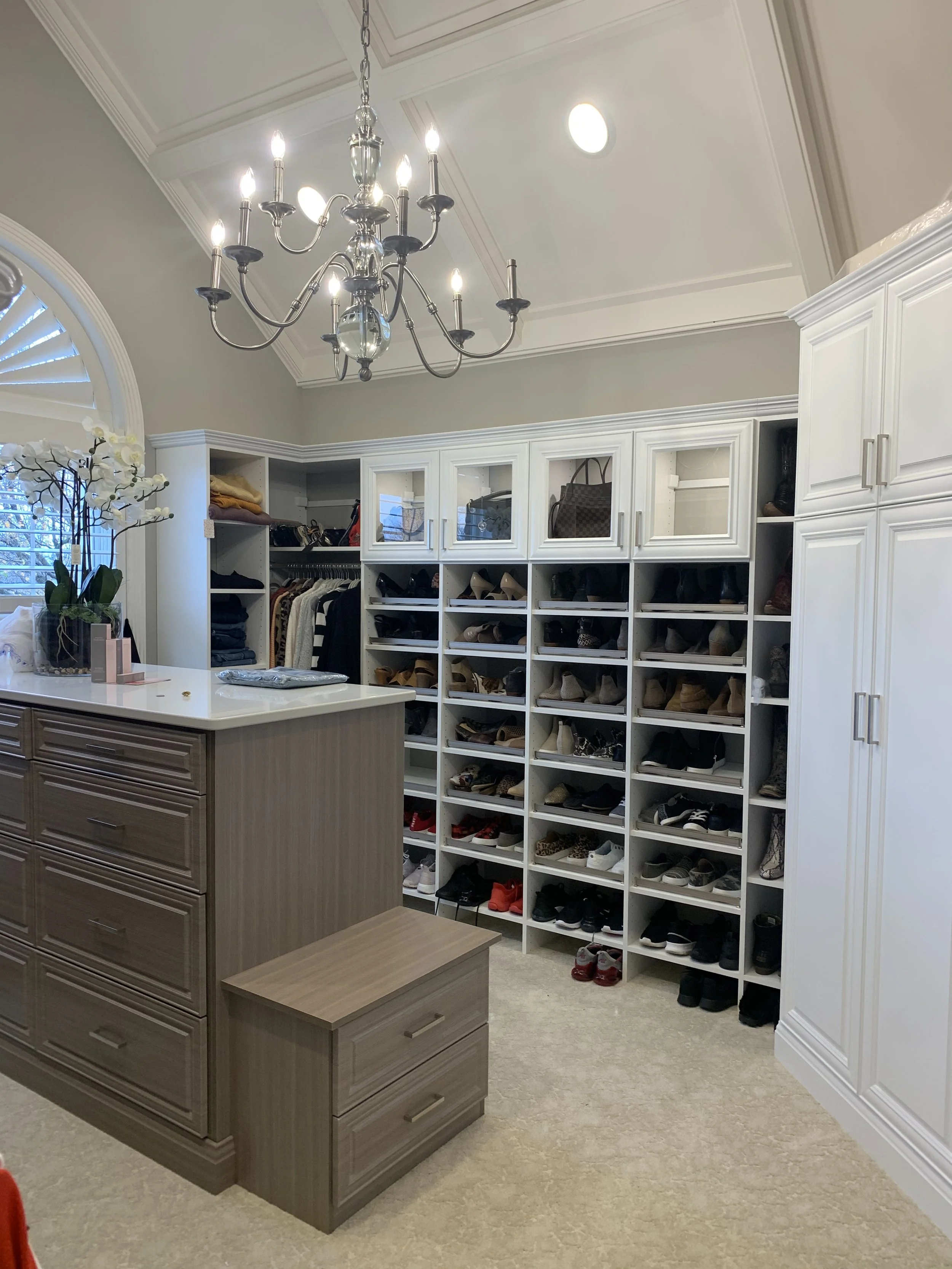 A walk-in closet with white cabinetry and shoe storage, a chandelier hanging from the ceiling, a window with blinds, and a white island with drawers and a small set of stairs.