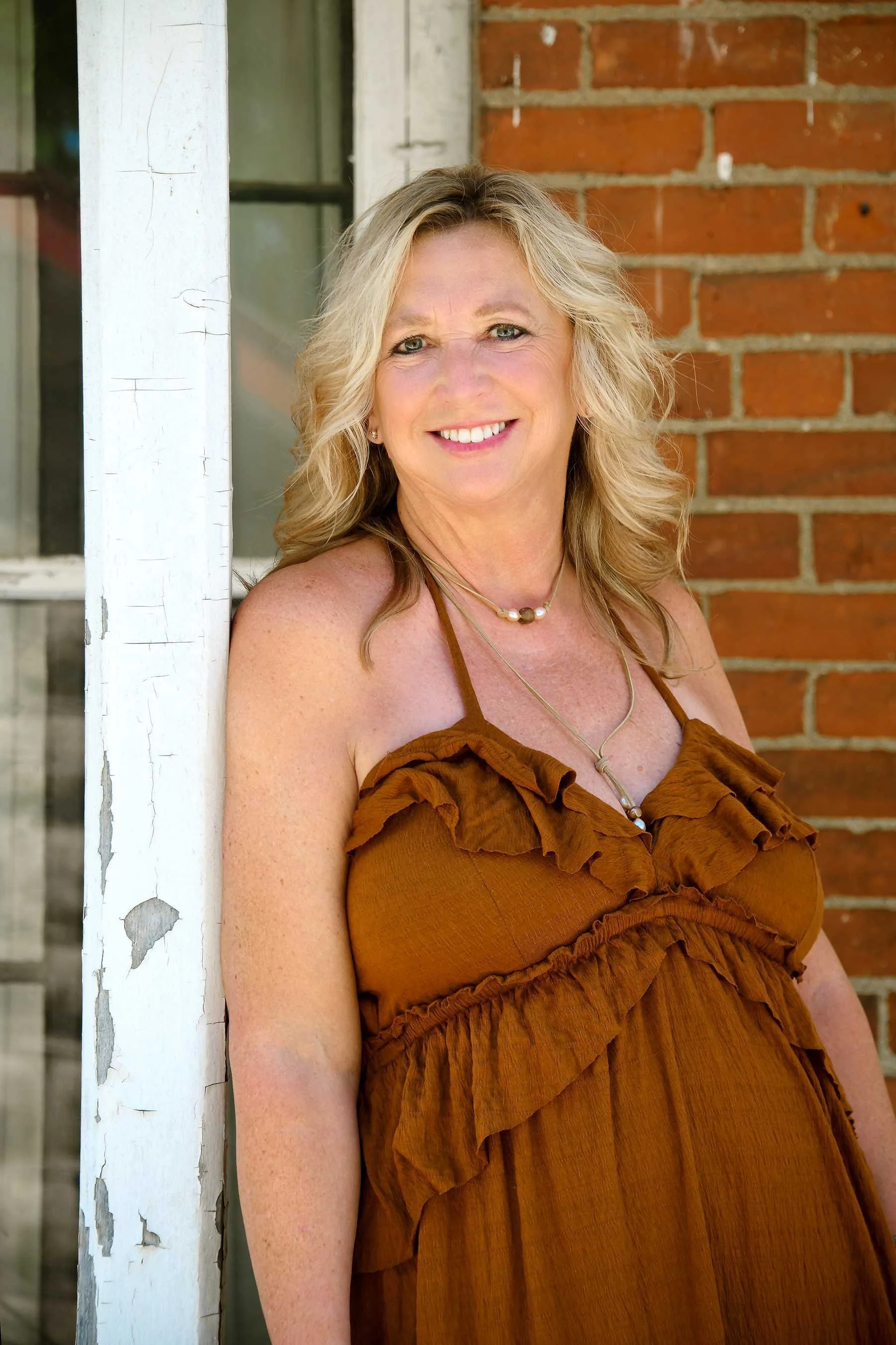 A smiling woman with blonde curly hair leans against a white weathered post in front of a brick building, wearing an orange ruffled dress and jewelry.