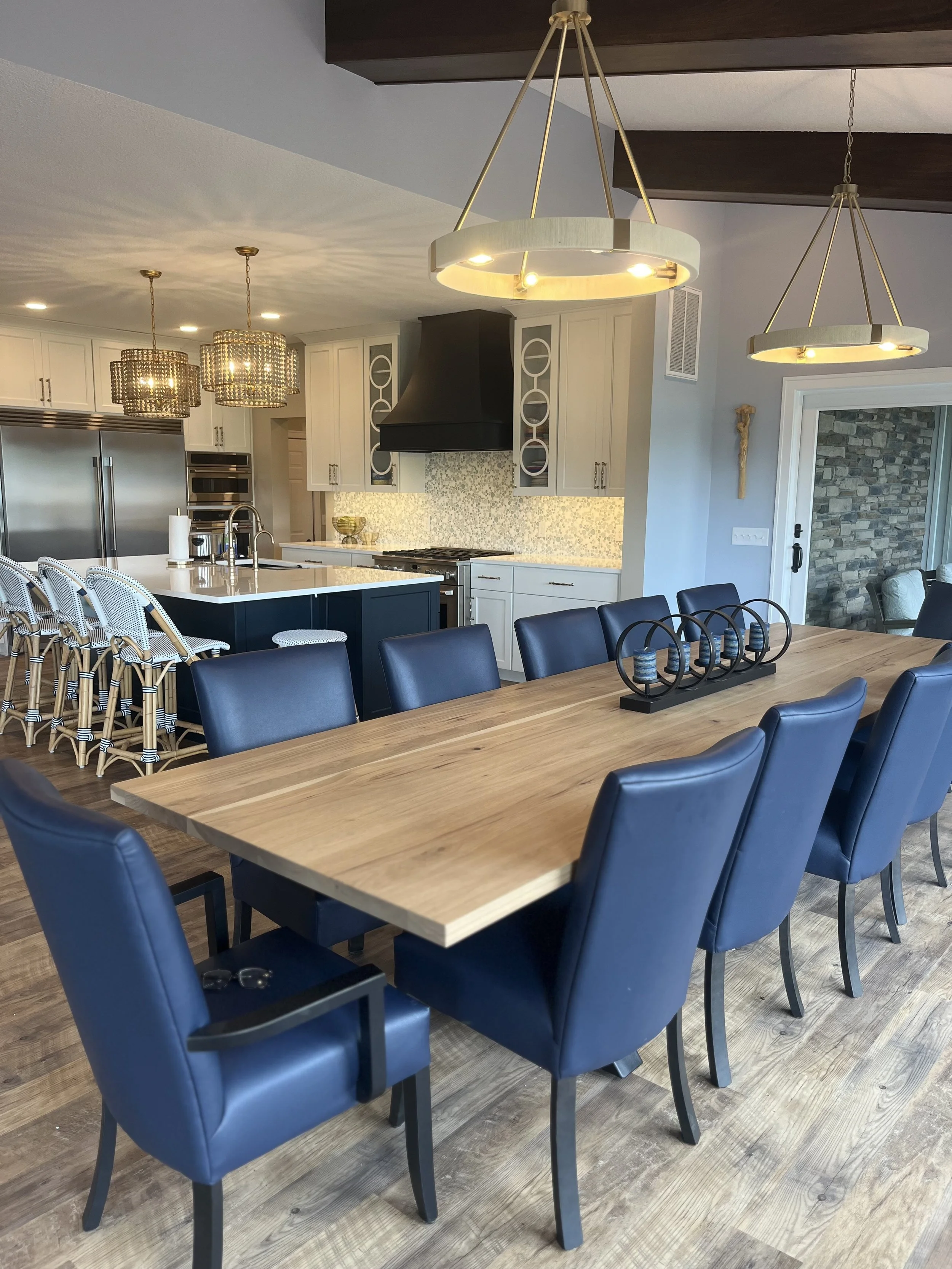 Modern kitchen and dining area with a large wooden table surrounded by eight blue leather chairs. The kitchen features white cabinets, a black range hood, a mosaic tile backsplash, and stainless steel appliances. Two pendant lights hang over the table, and three chandeliers are above the kitchen island with barstools. The flooring is wooden, and there is a sliding door with stone wall visible in the background.