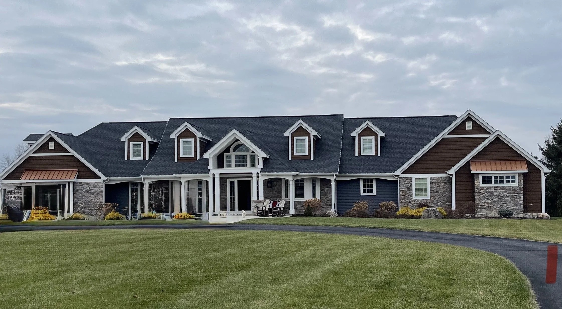 A large, modern house with a combination of stone and wooden siding, multiple gabled roofs with dark shingles, and several windows. The front yard has a curved driveway and well-maintained lawn with some plants and flowers.