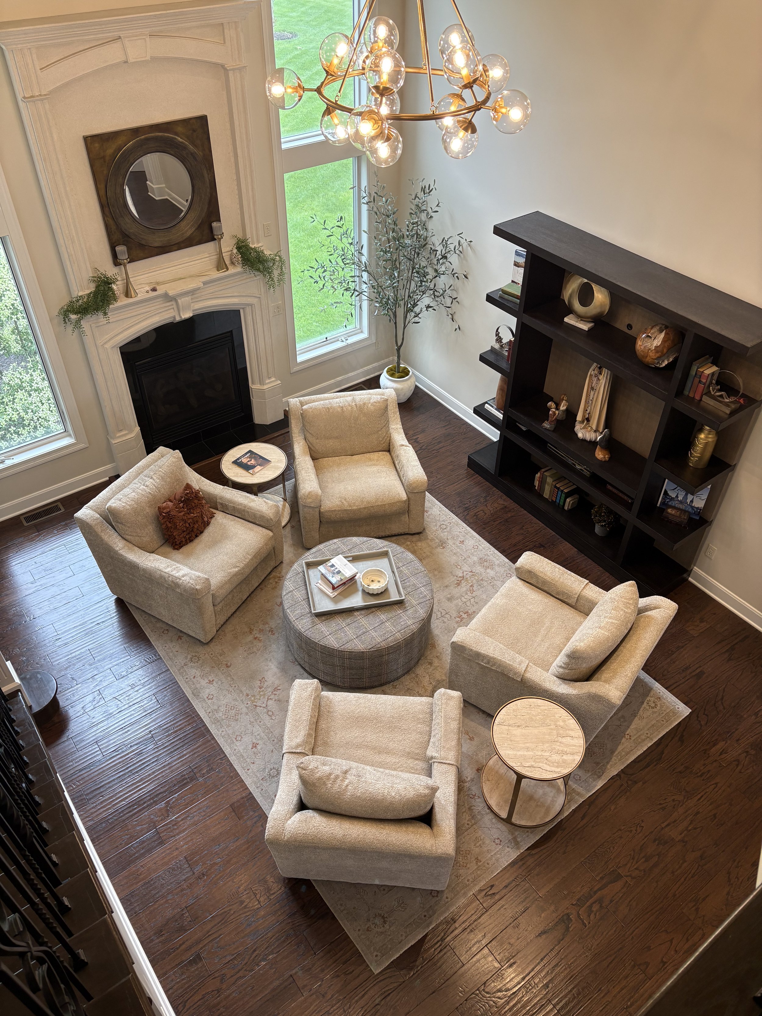 Living room with five beige armchairs arranged around a circular patterned ottoman on a beige area rug. There are two small round side tables, one with books and a candle. A black bookshelf with decorative items stands against the white wall, and a fireplace with a large round mirror and greenery decor above it. Two large windows let in natural light, with a tall potted plant next to the windows. A modern chandelier with exposed bulbs hangs from the ceiling.