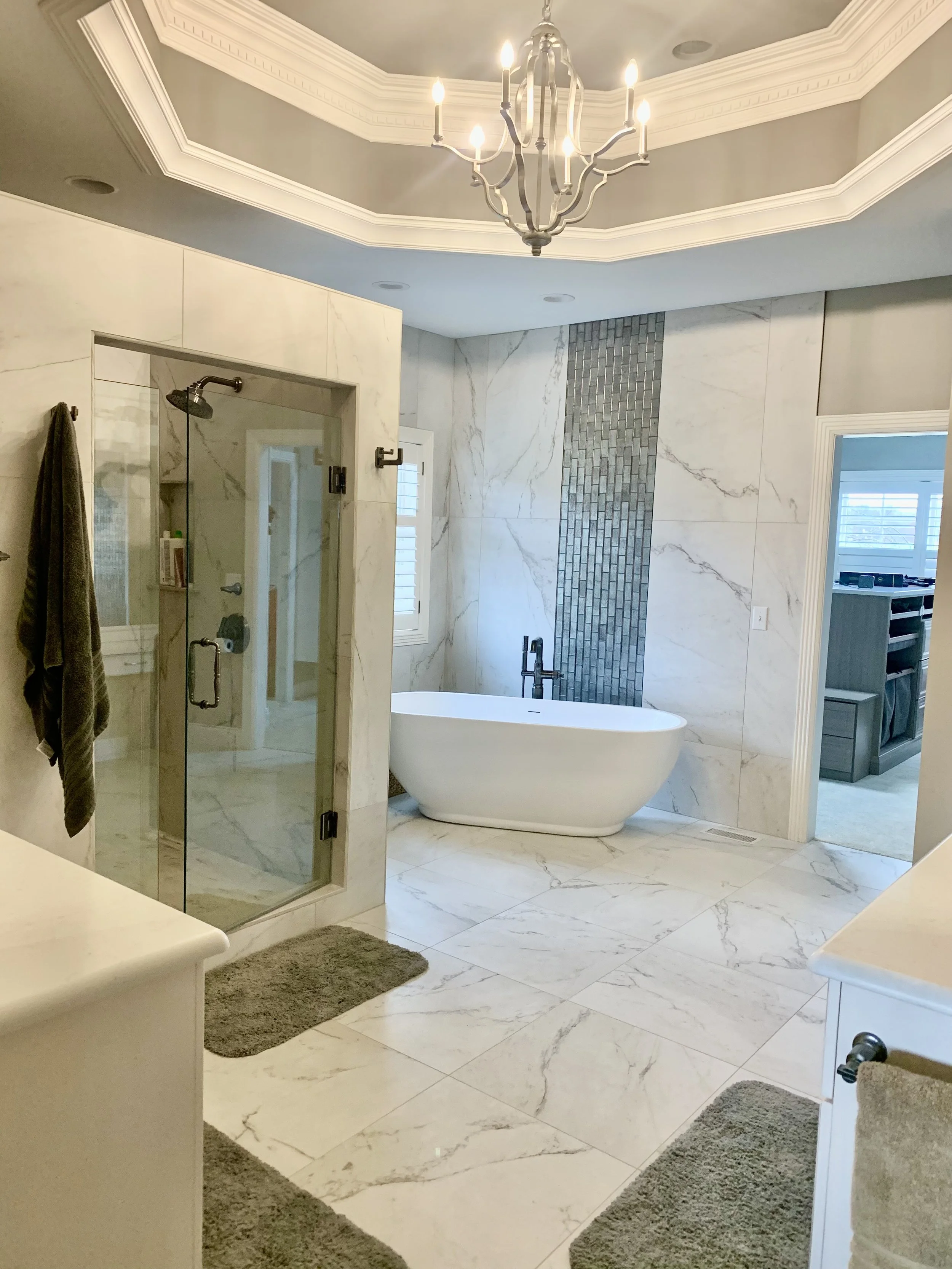 A luxurious bathroom featuring a glass-enclosed shower, a freestanding bathtub with a black faucet, marble walls and flooring, a chandelier overhead, and a window with white shutters.