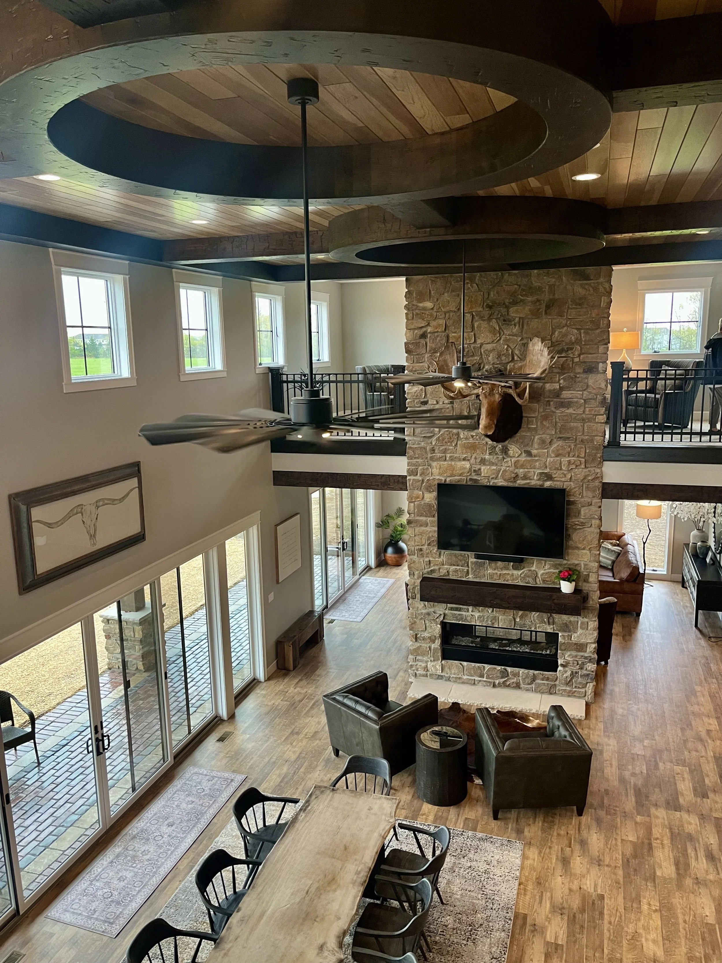 A spacious living room with a high ceiling, wooden accents, and large windows. There is a stone fireplace with a mounted TV above it, seating area with leather chairs, a dining table with chairs, and a balcony with black railing.