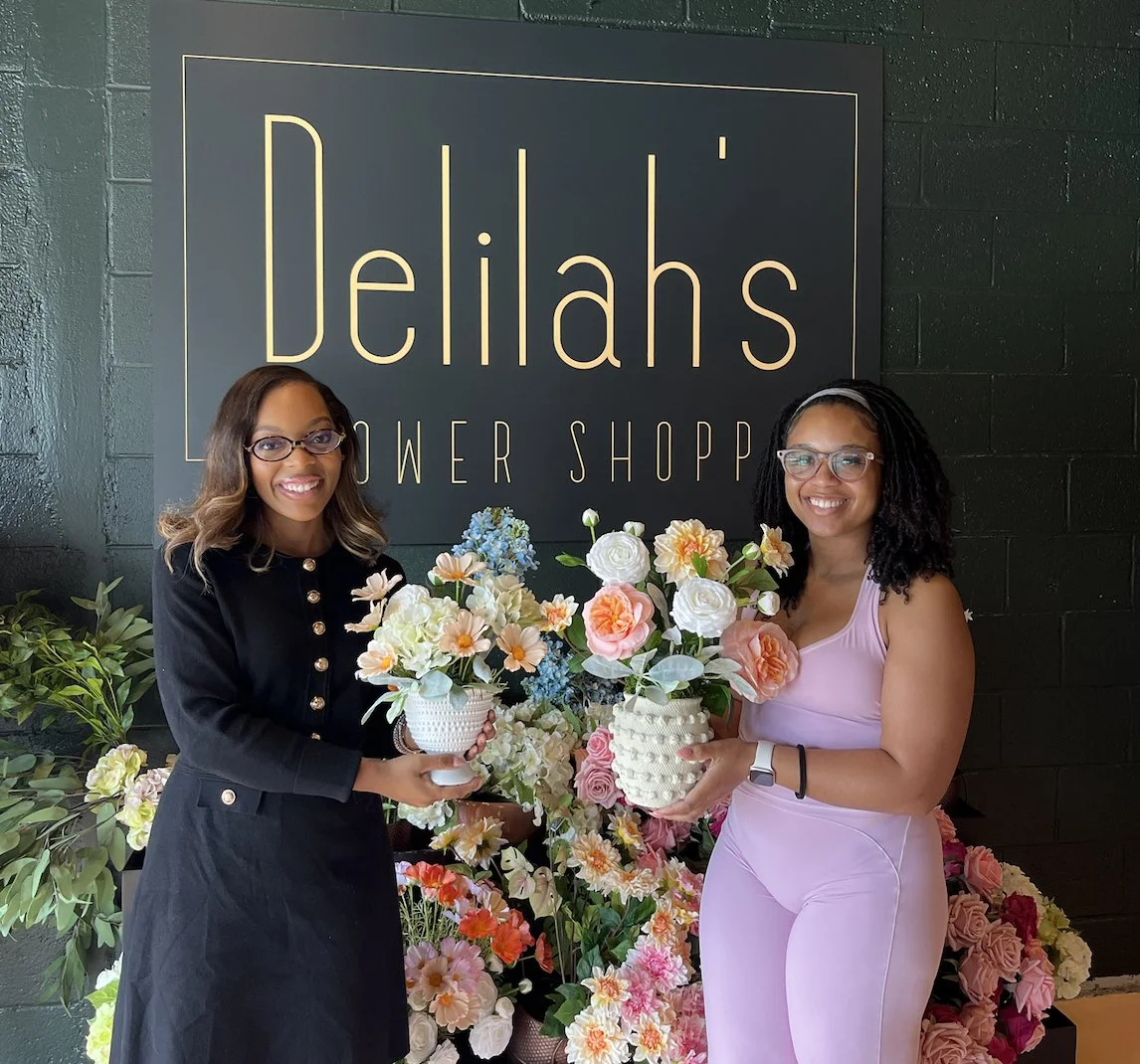 Two women smiling and holding flower arrangements in front of a sign that says "Delilah's Flower Shop" with an array of flowers behind them after completing a bouquet making class.