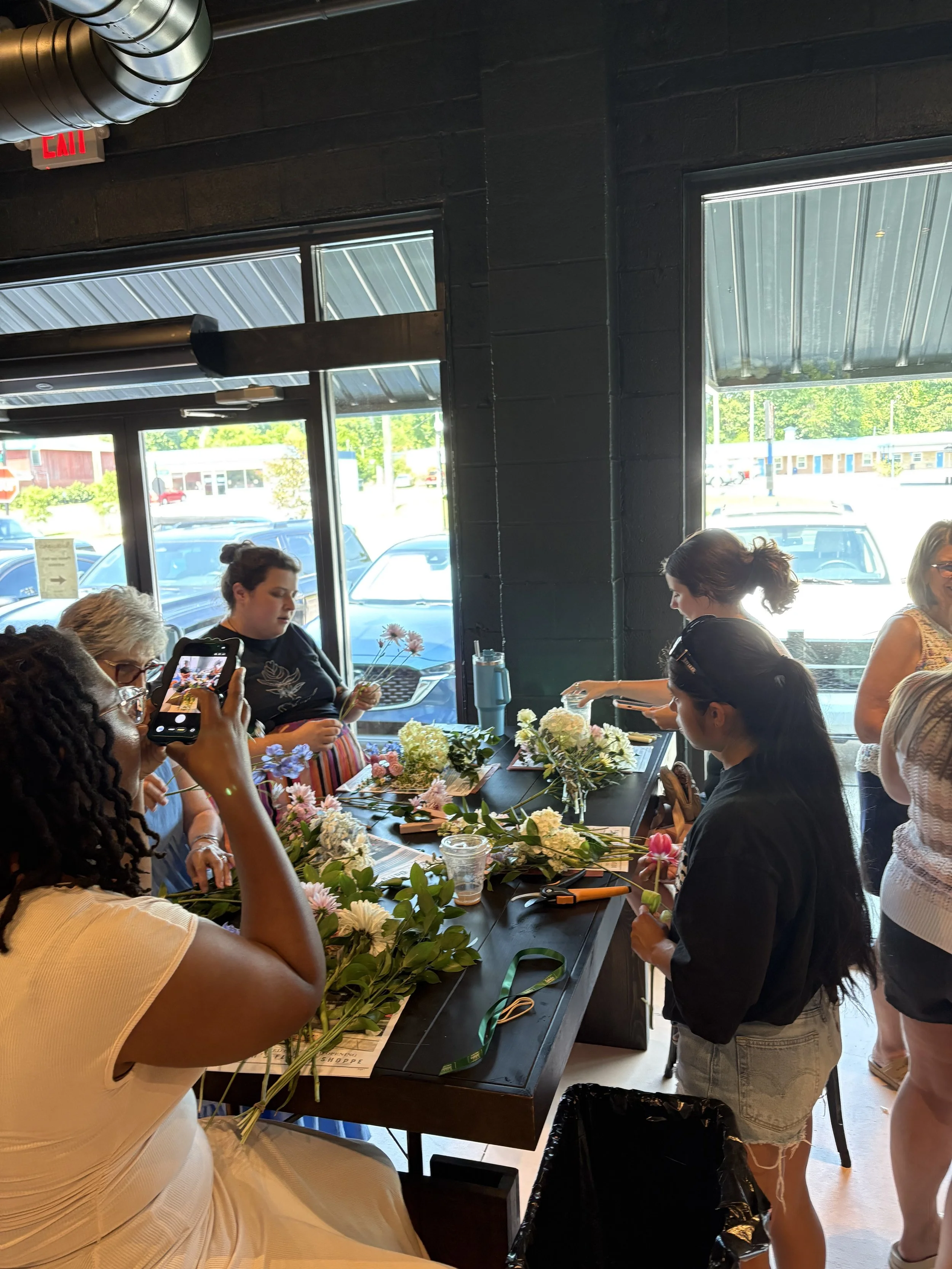 Group of women making flower arrangements at a table during a bouquet workshop class, with large windows showing parked cars outside.