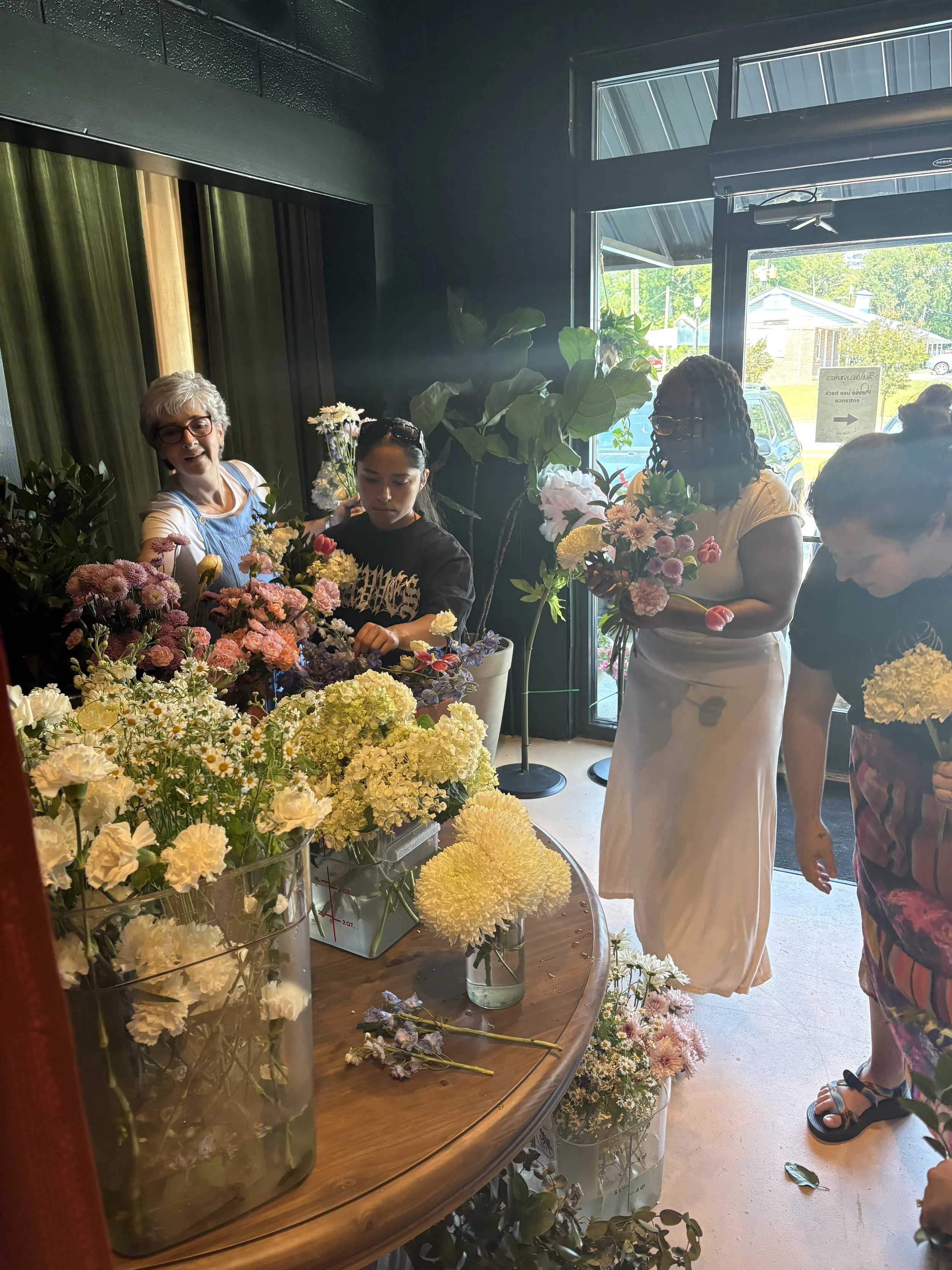 People arranging and selecting various colorful flowers in a flower shop with large windows and natural daylight.