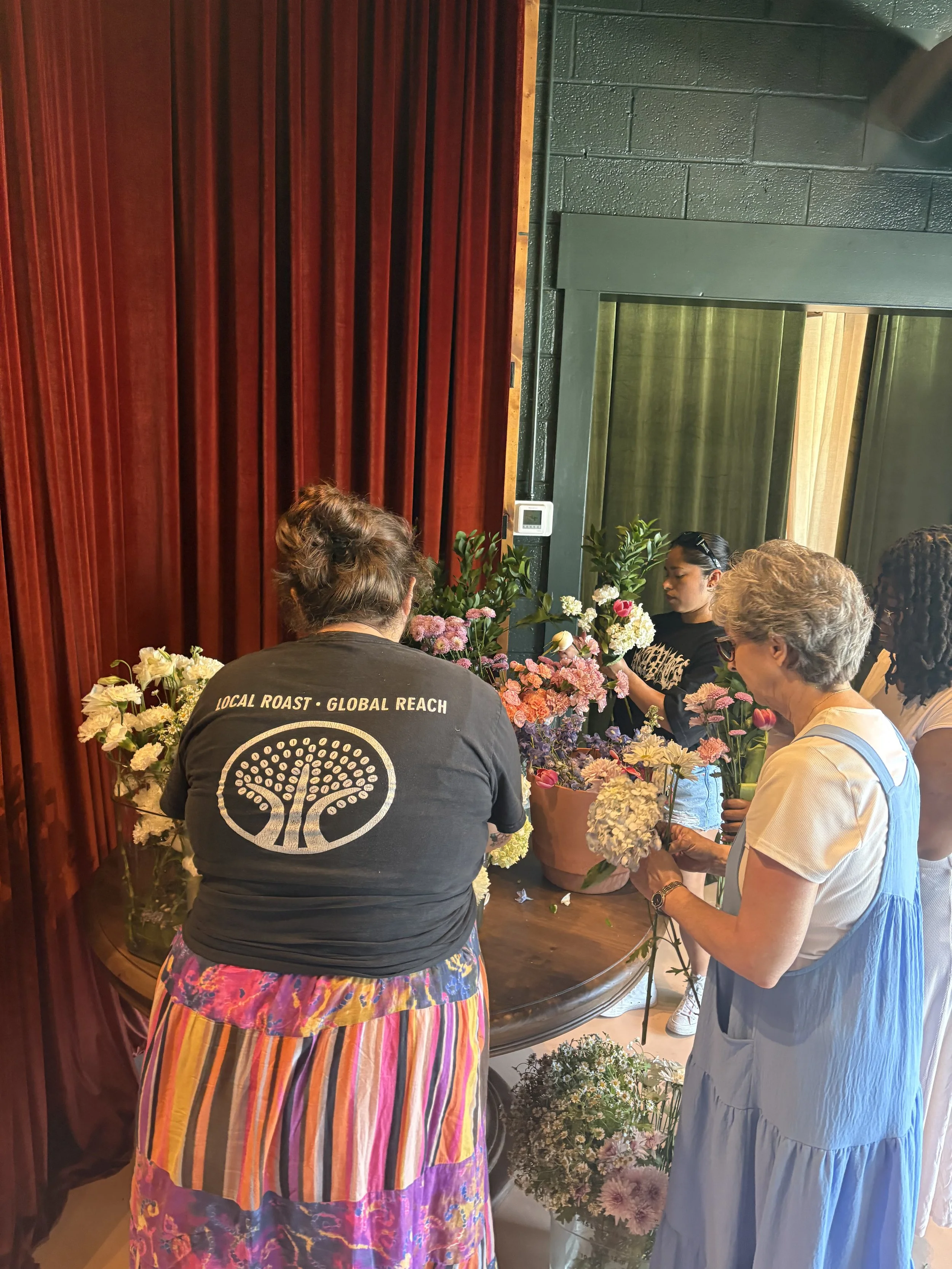 Multiple women gathered around flowers, arranging and holding flower bouquets indoors, with a dark green wall, a window with curtains, and wooden paneling in the background.