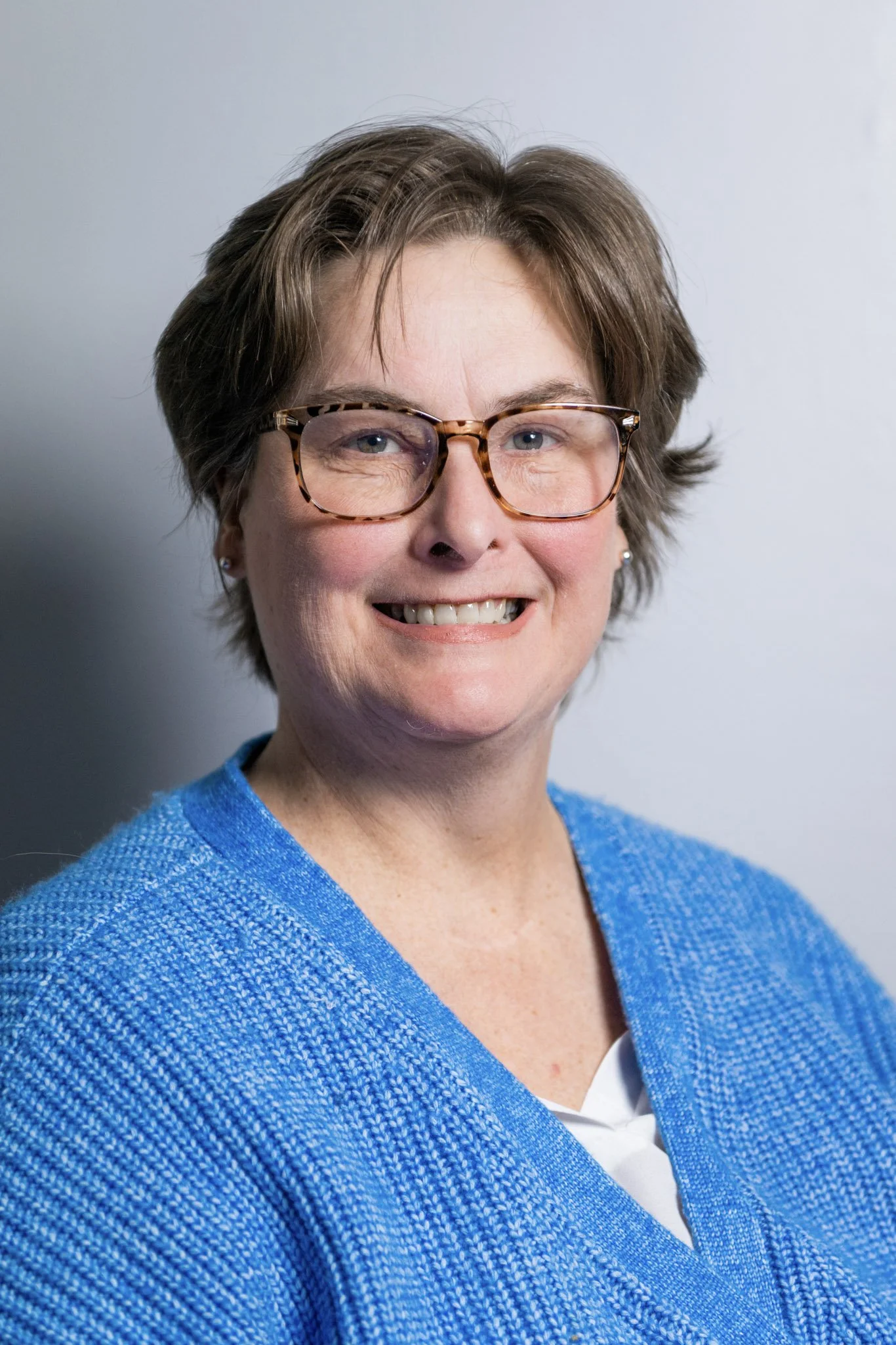 A smiling woman with short brown hair, glasses, and earrings, wearing a blue textured cardigan over a white top, posing against a plain gray background.