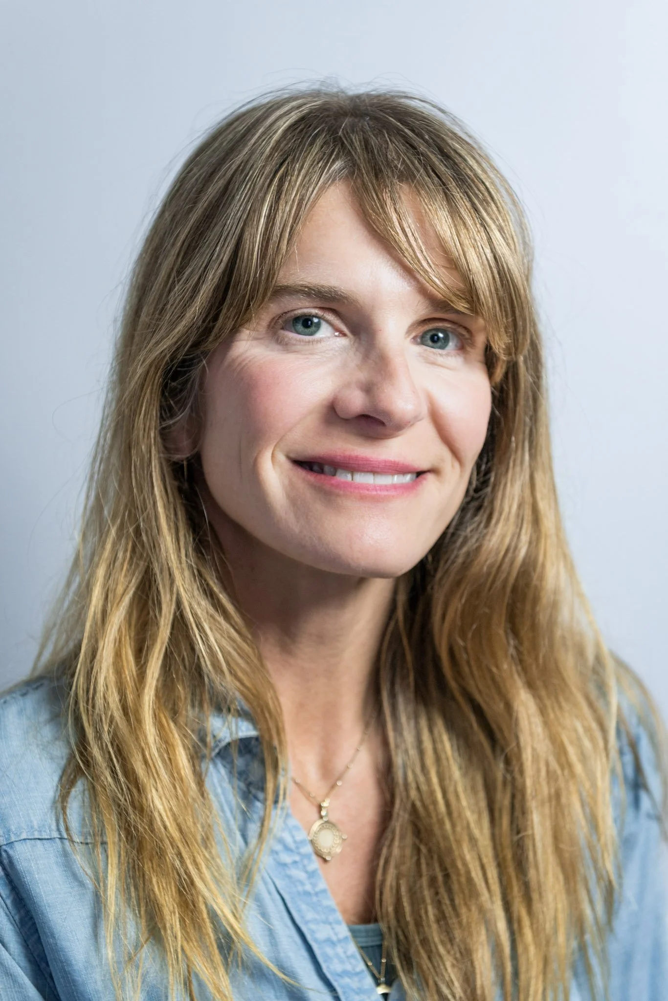 Close-up portrait of a woman with long, light brown hair, wearing a blue top and a gold necklace with a pendant, smiling softly against a plain background.