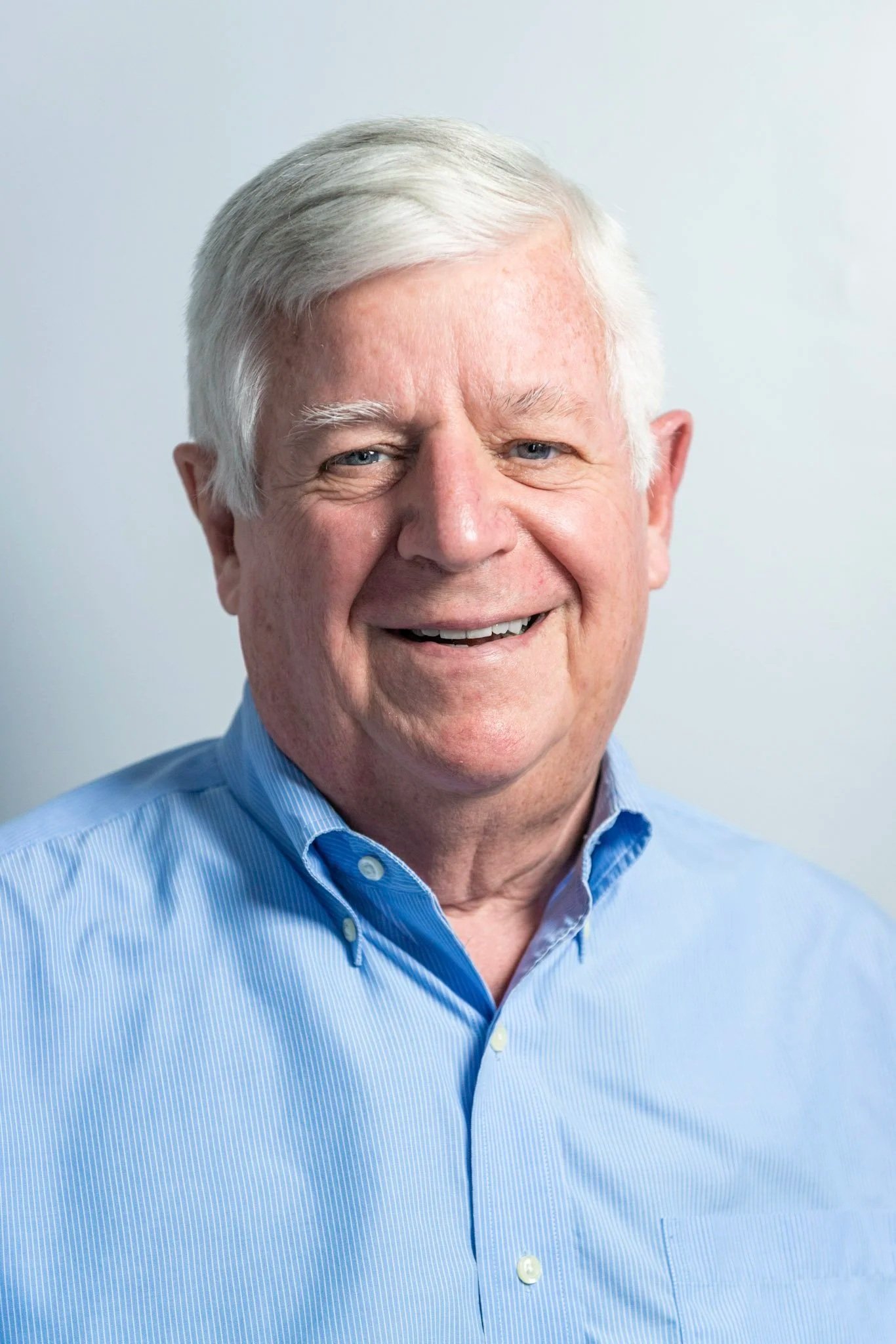 A smiling elderly man with gray hair wearing a pink checkered shirt.
