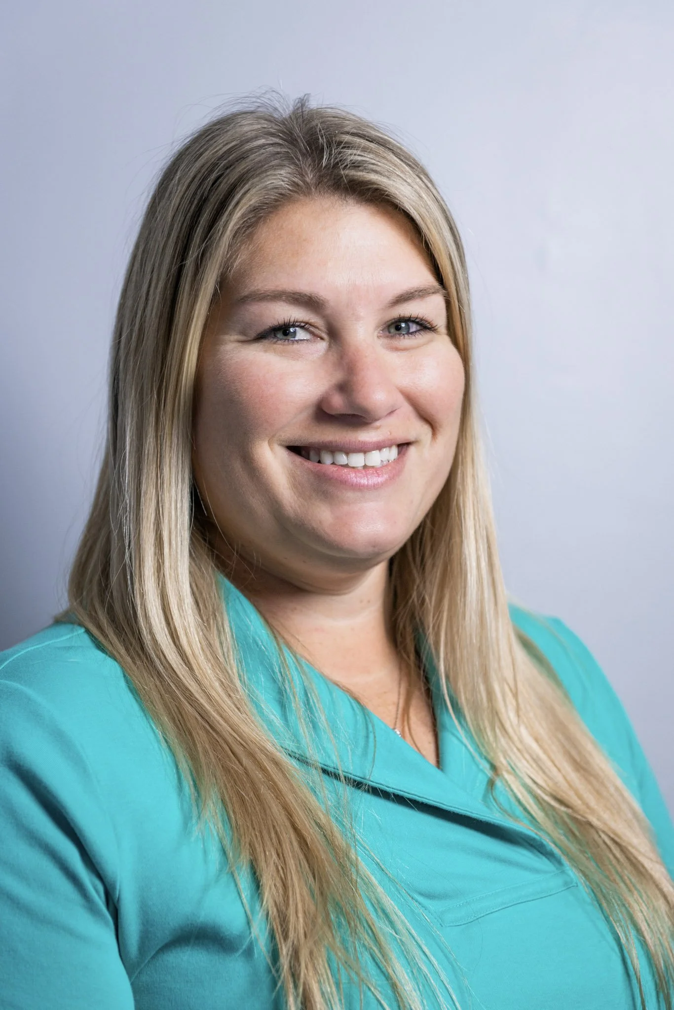 A smiling woman with long blonde hair wearing a turquoise top against a plain light background.
