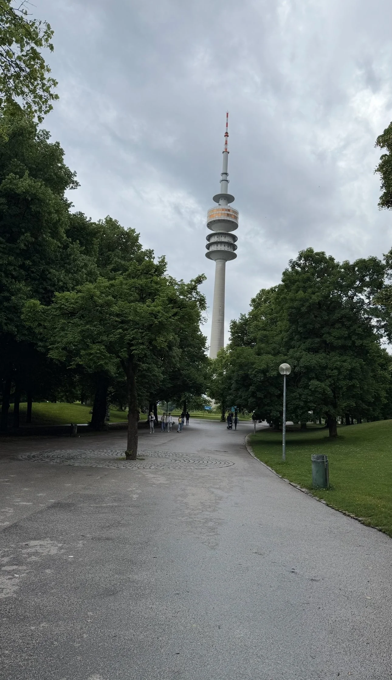 A park with a paved walkway, surrounded by green trees, with a tall television tower in the background under a cloudy sky.