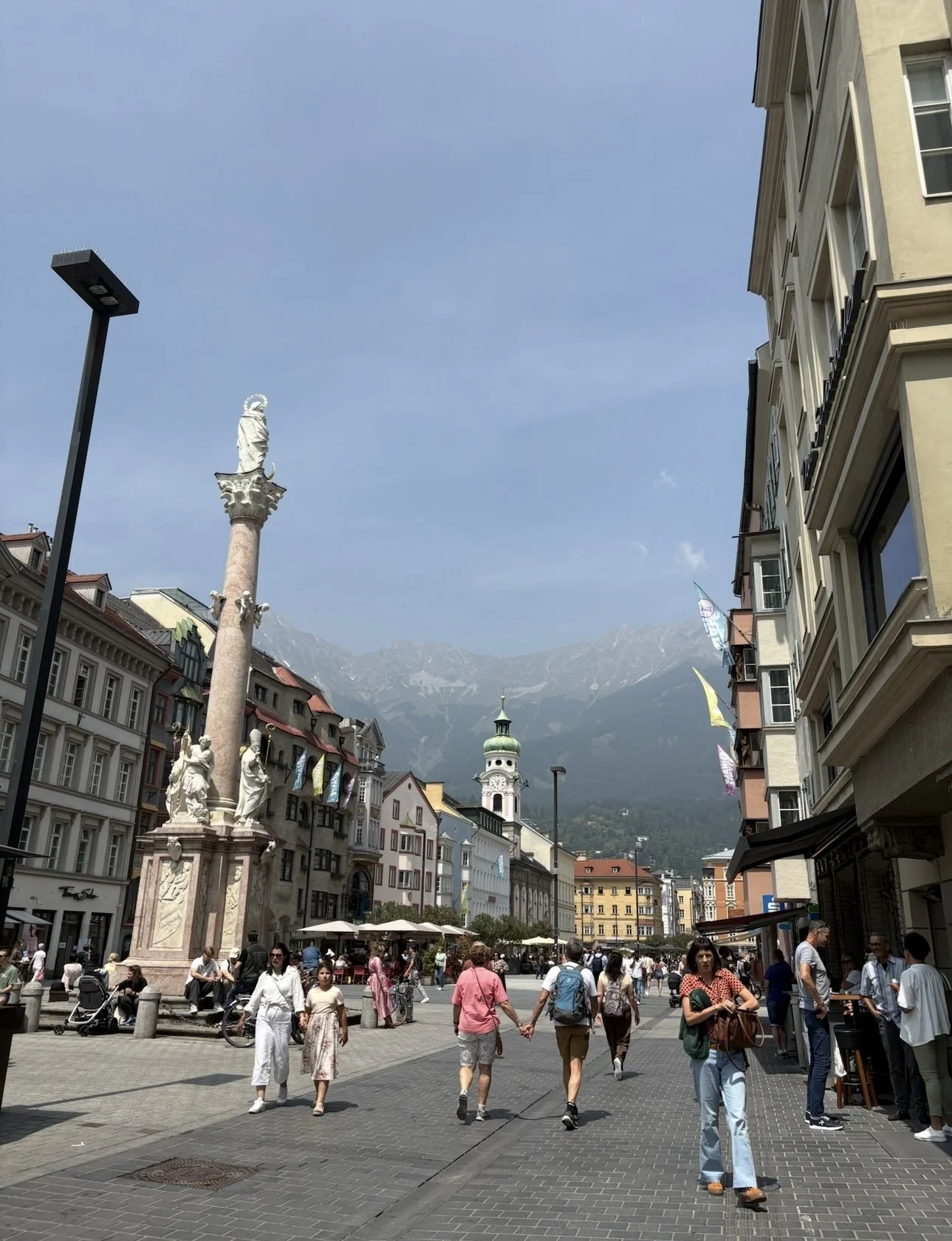 People walking on a busy European city street with historic buildings, a tall statue on a column, and mountains in the background under a blue sky.