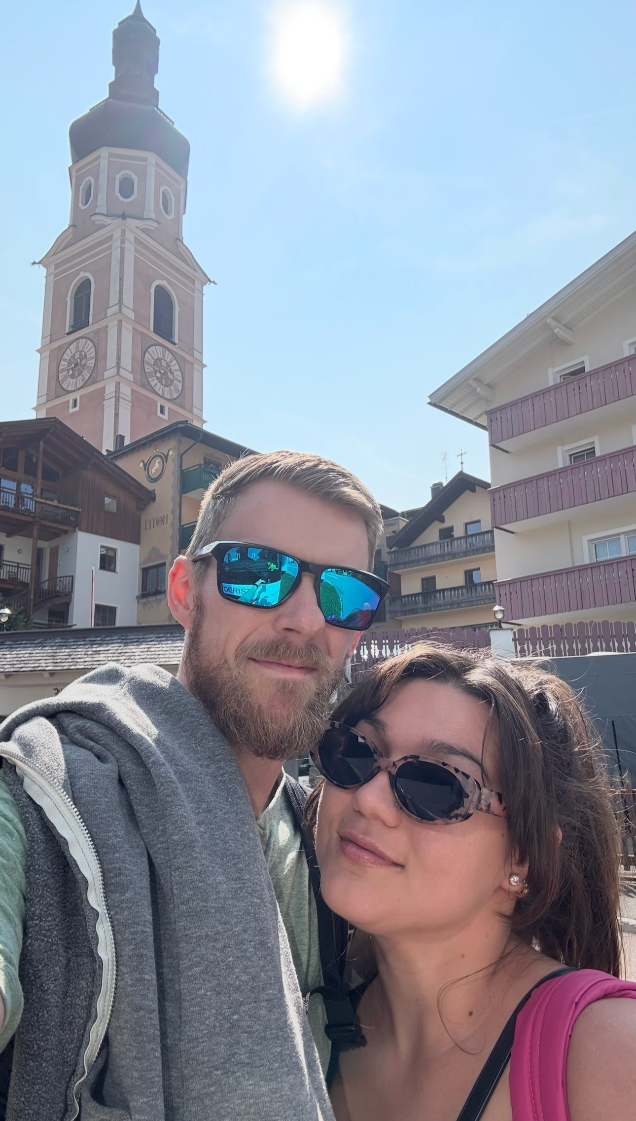 A man and woman taking a selfie outside on a sunny day. The man has short hair, a beard, and is wearing blue sunglasses and a gray hoodie. The woman has long dark hair, sunglasses, and a pink top. In the background, there is a tall clock tower and buildings that resemble a European village.