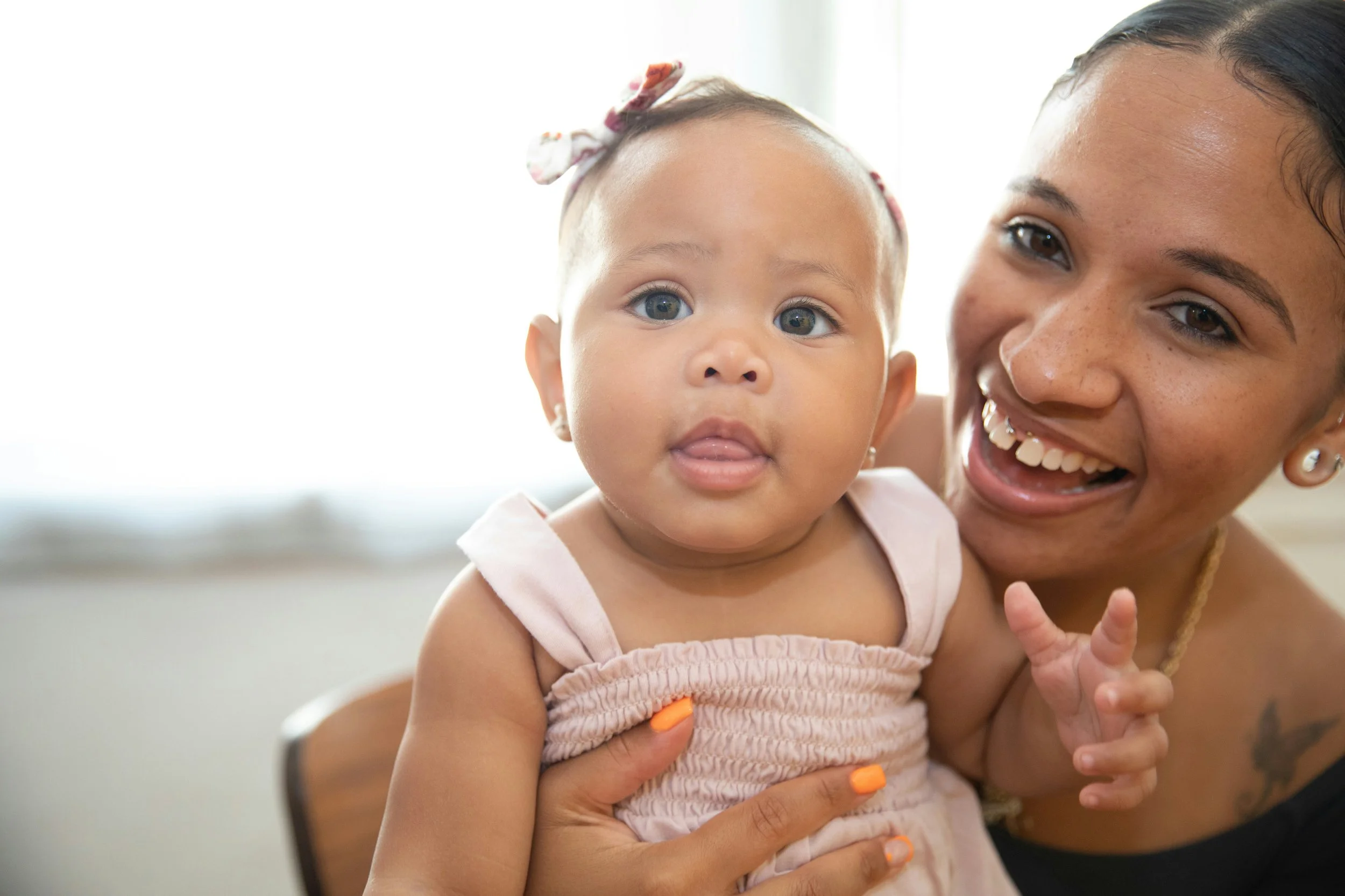 A smiling woman holding a young girl with curly hair, wearing a pink dress and a bow in her hair, in a bright room.