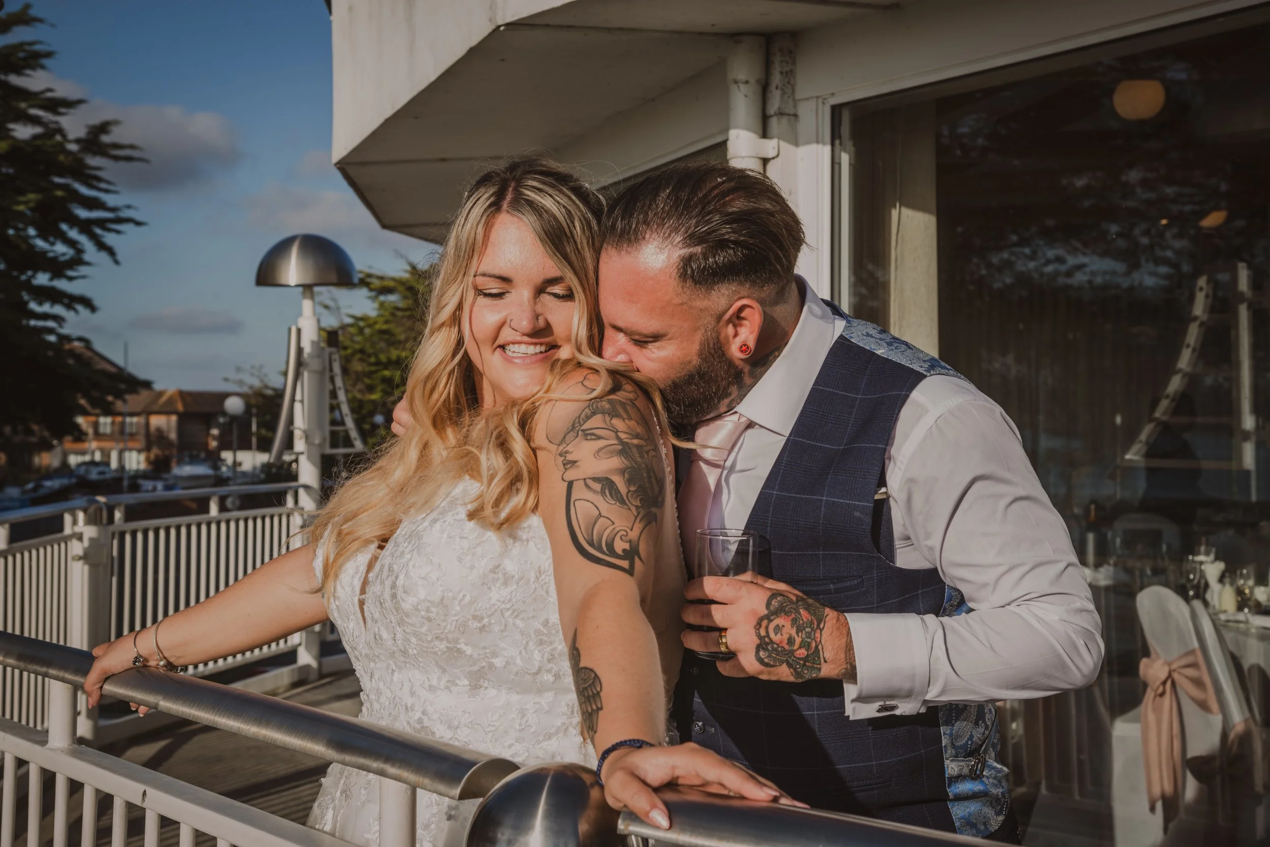 A smiling woman in a white lace dress and a man in a blue vest and white shirt sharing an intimate moment on an outdoor balcony during daytime.