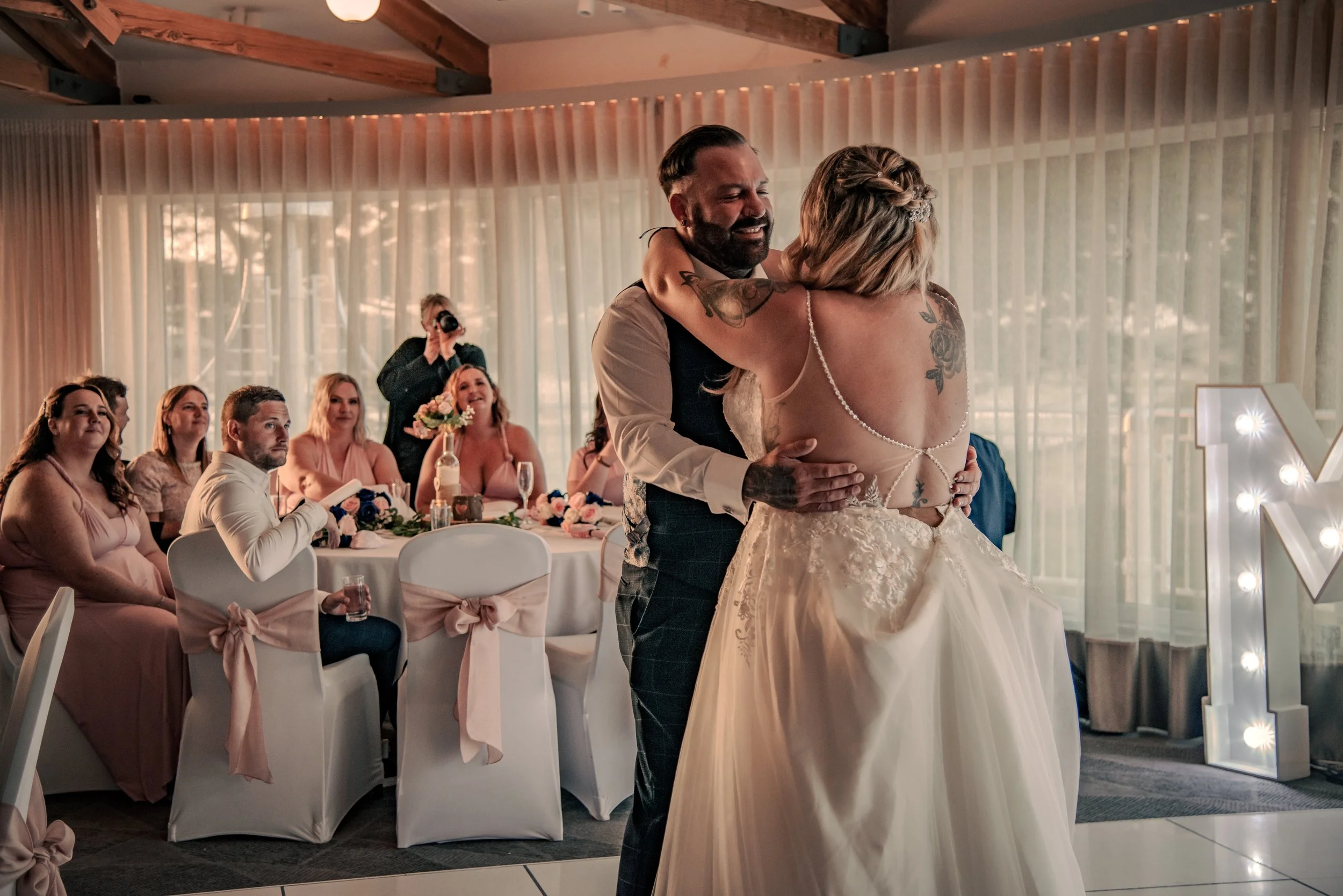 A bride and groom sharing a dance at their wedding reception, with guests seated at decorated tables in the background, inside a decorated venue with warm lighting.