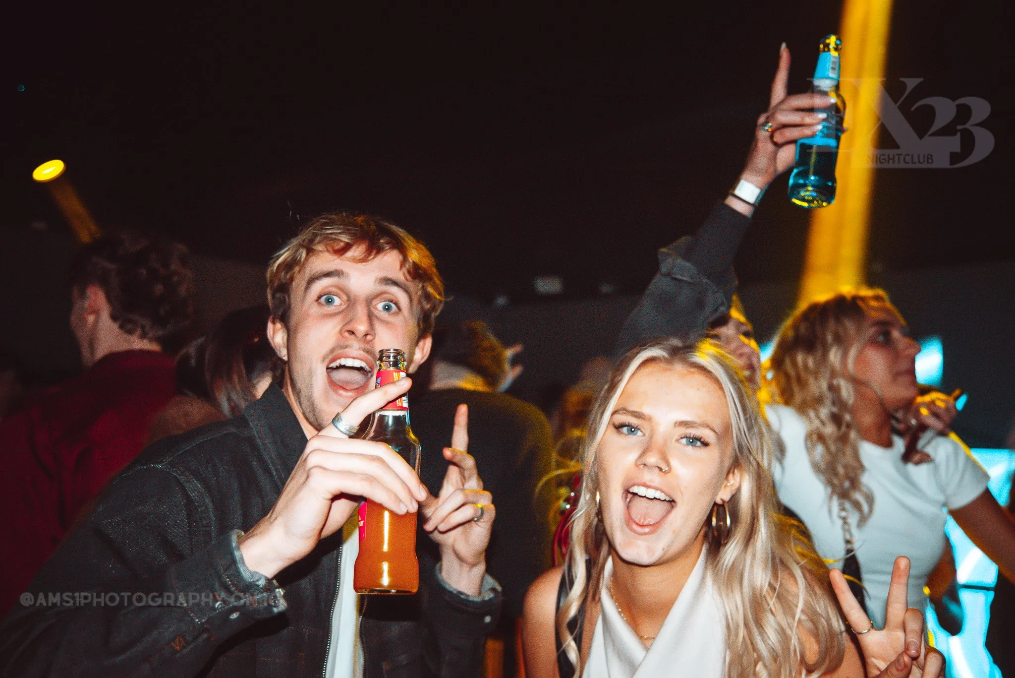 People dancing and celebrating at a nightclub, with two individuals in the foreground holding drinks and making peace signs.