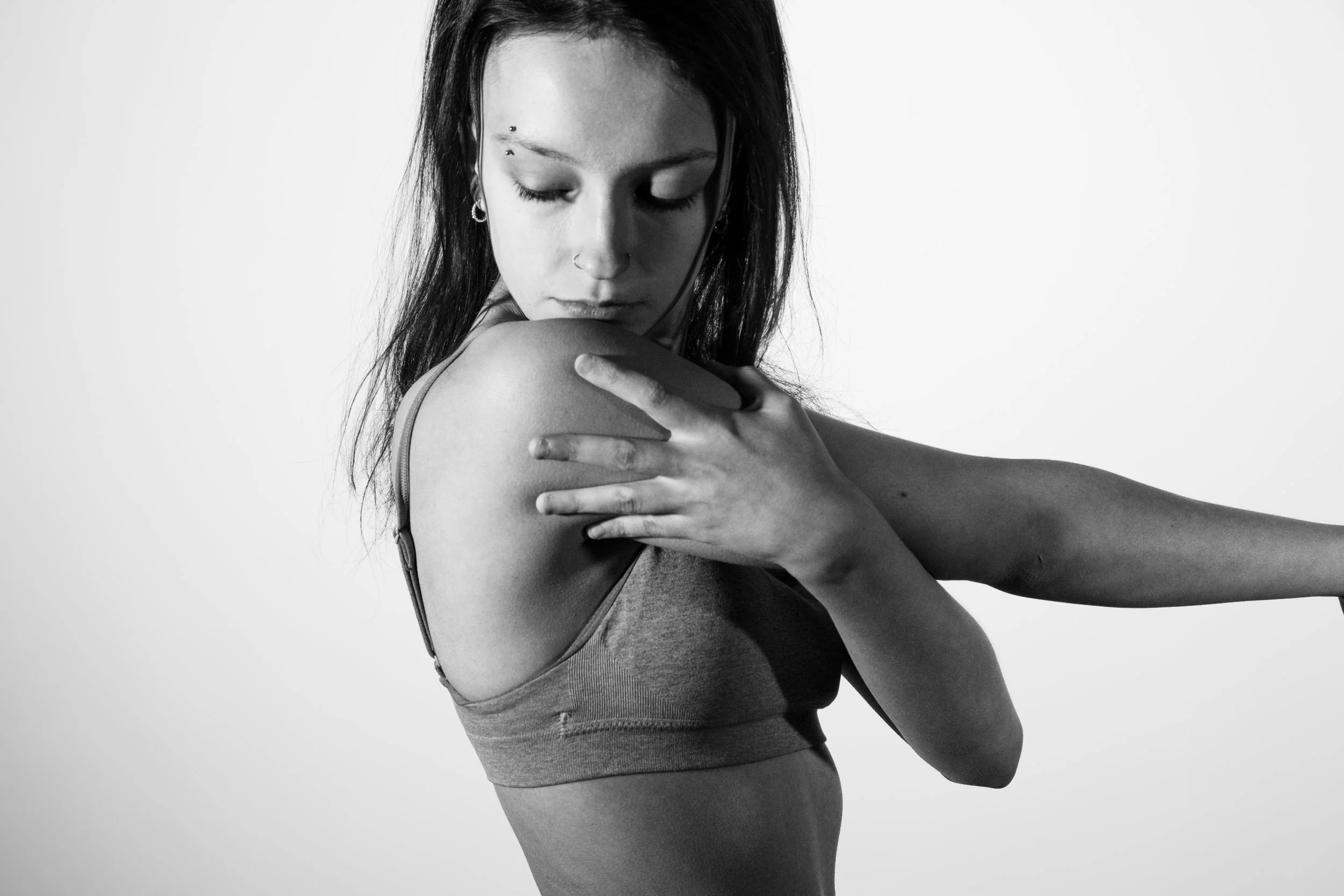 Black and white photo of a young woman with dark hair, wearing a sports bra, touching her shoulder and looking down with her eyes closed.