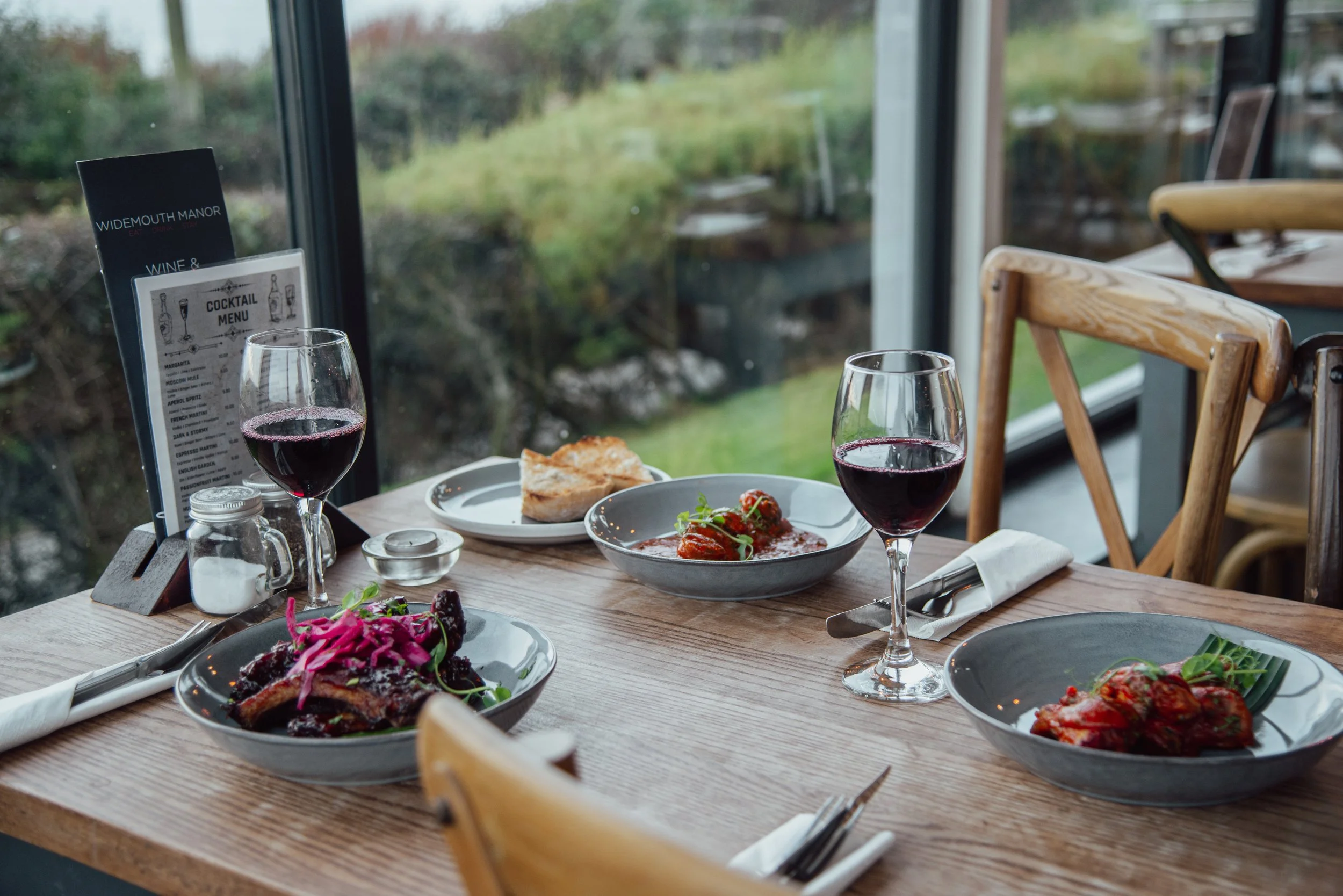A wooden table set with two glasses of red wine, two plates with meat and vegetables, a slice of bread, a salt shaker, a small container, and a cocktail menu, all near a large window overlooking a garden.