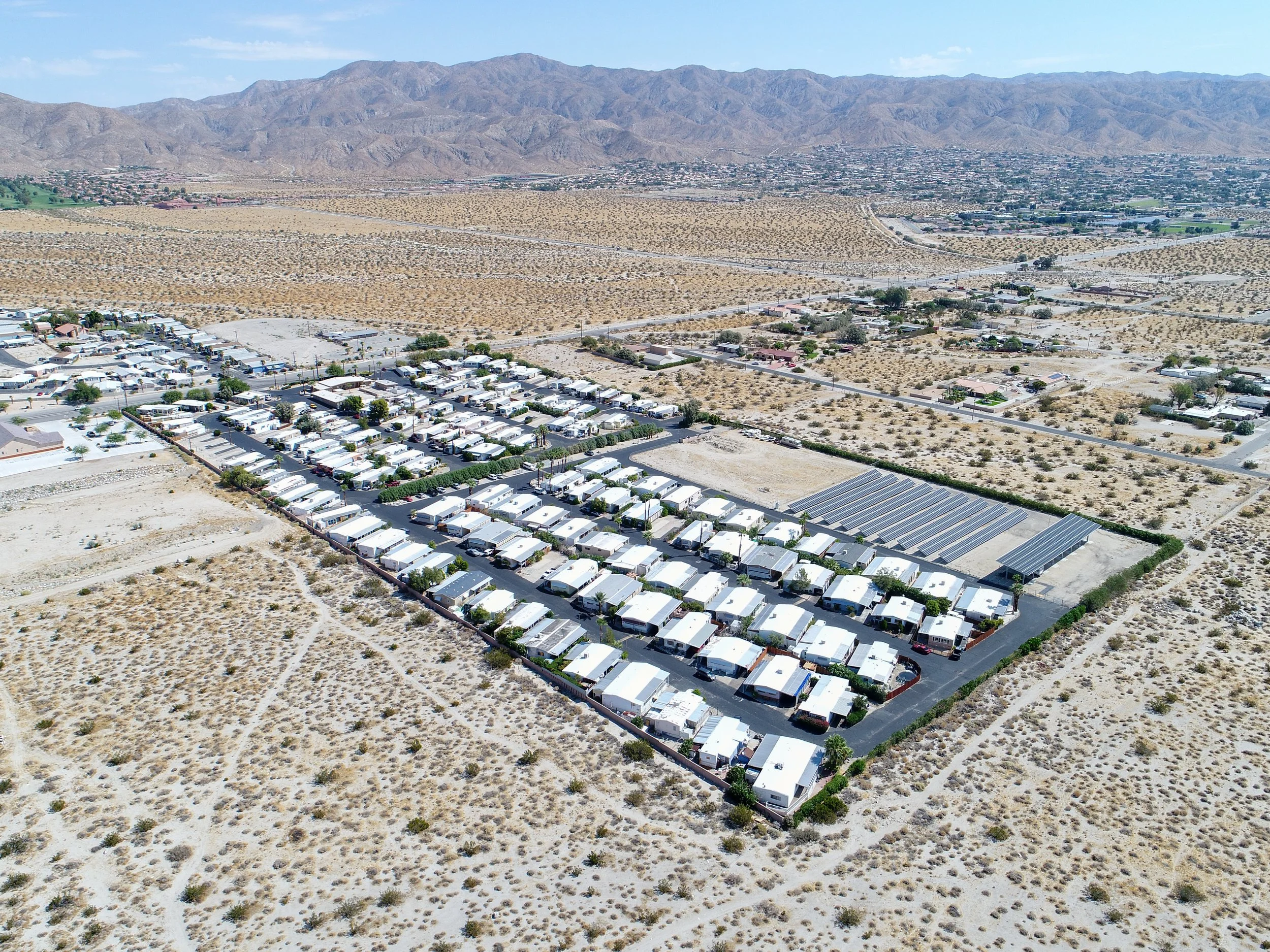 Aerial view of a residential area in a desert landscape with mountains in the background. The area contains rows of mobile homes and solar panels, surrounded by dry vegetation and sparse development.