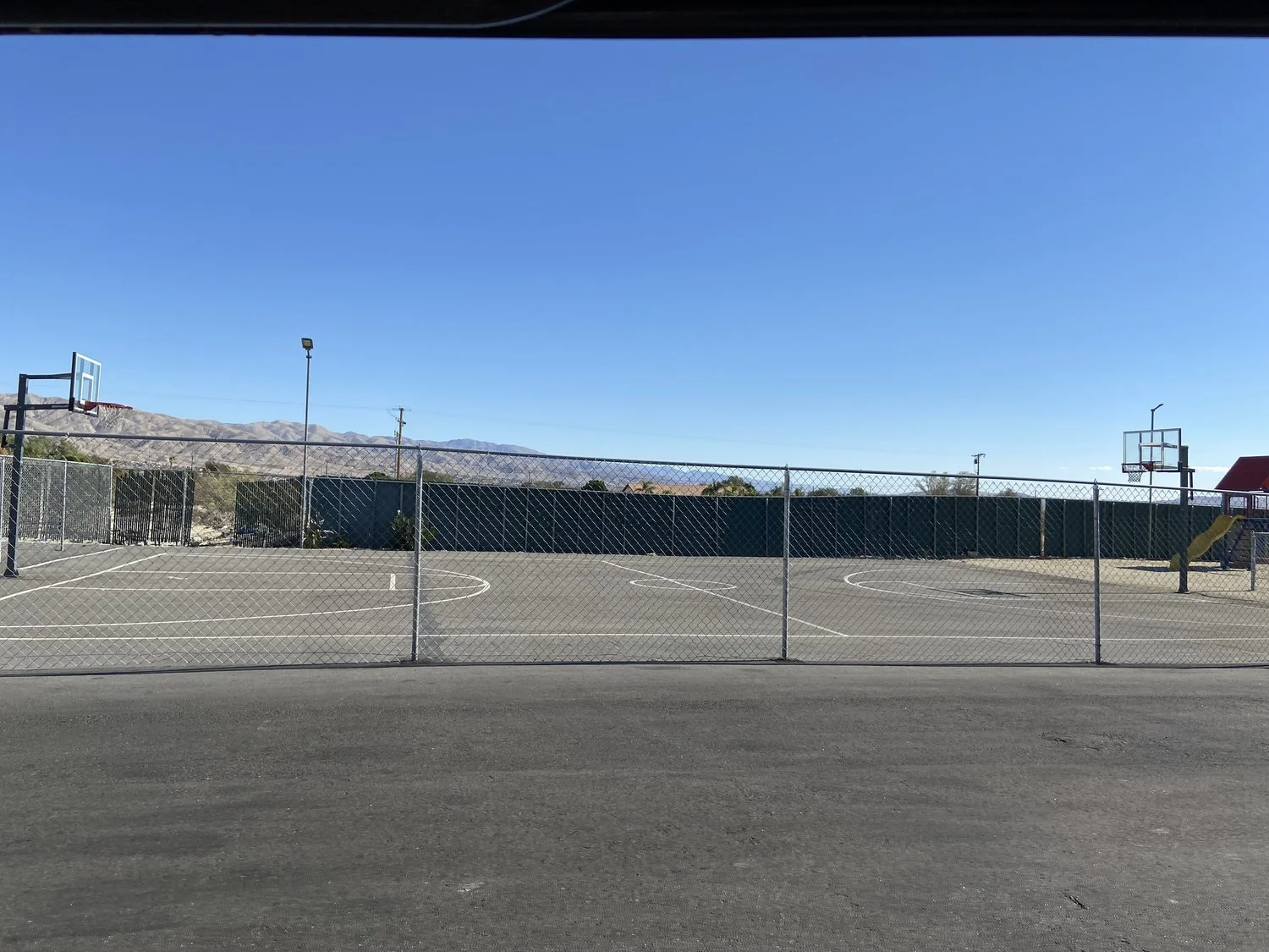 Empty outdoor basketball court with chain-link fence, two hoops, and mountain landscape in the background.