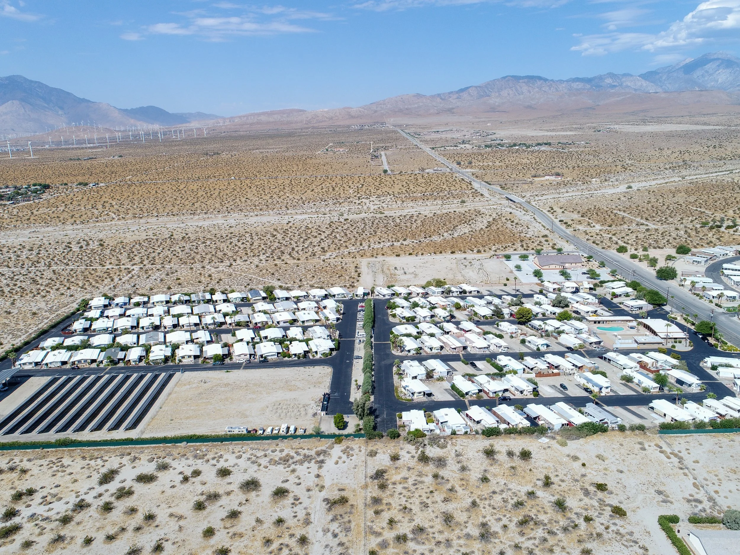 Aerial view of a desert landscape with a mobile home park and surrounding roads, set against a backdrop of mountains and wind turbines.