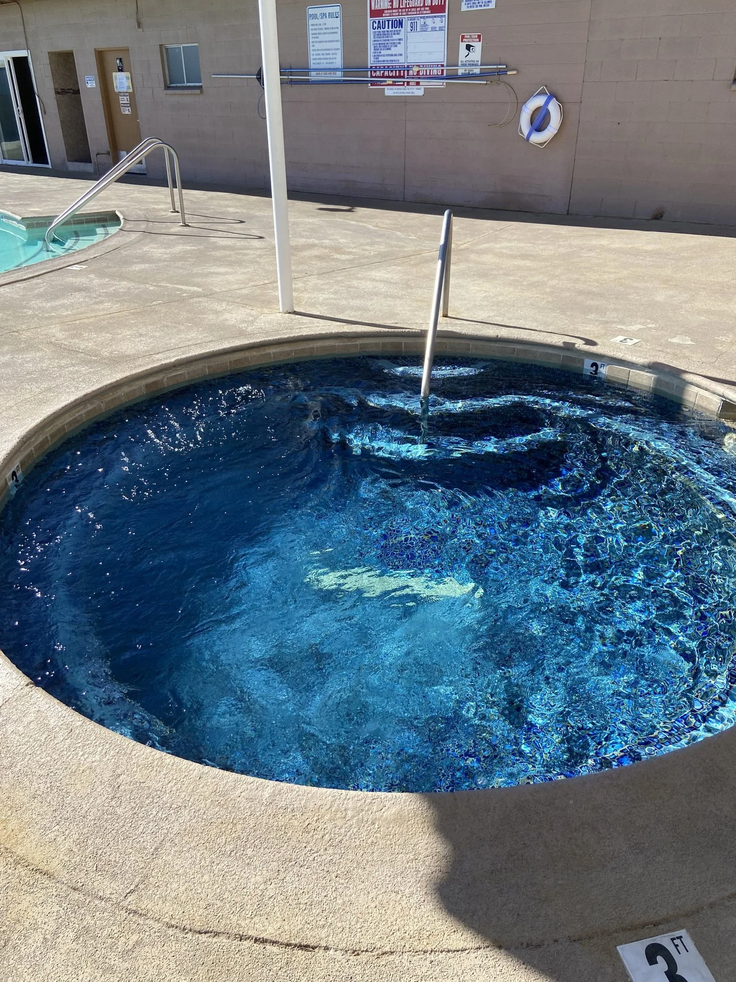 Circular hot tub with clear blue water next to a pool, surrounded by a concrete deck. Safety signs and a life preserver are visible on a nearby wall. A metal handrail provides access to the hot tub.