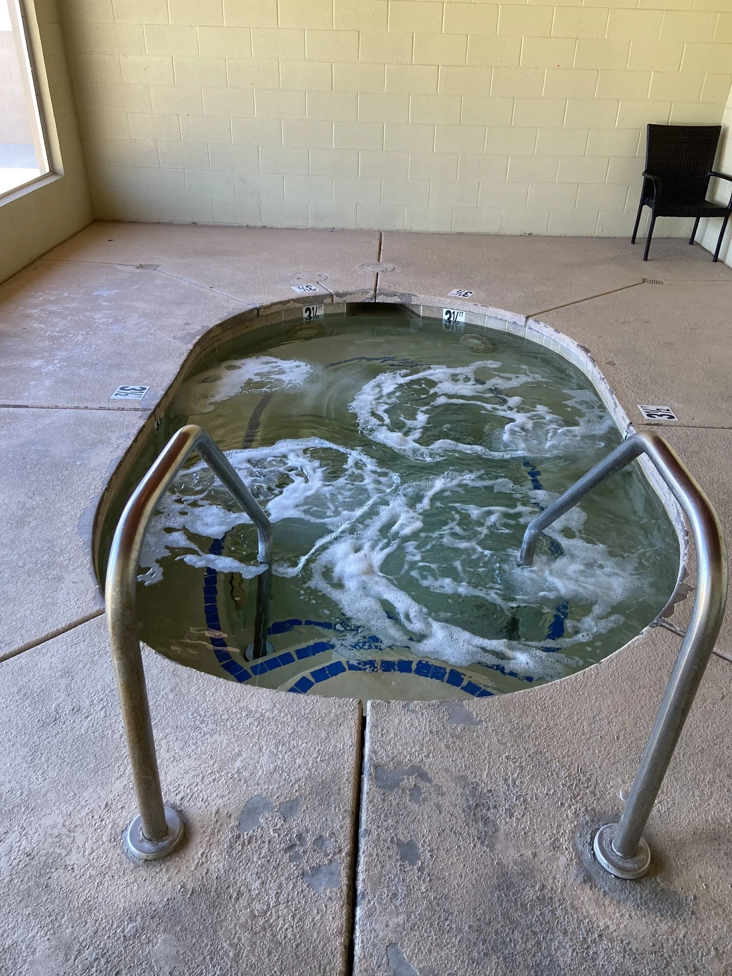 Small indoor hot tub with bubbling water, surrounded by a tiled edge and metal handrails, with a chair in the corner.