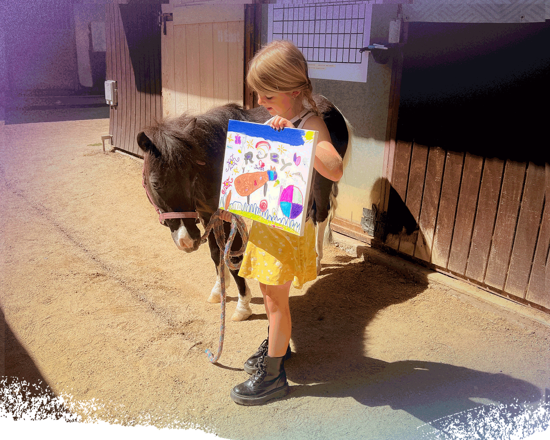 A young girl with long braids wearing a yellow skirt, holding a colorful drawing board, and standing next to a small black and white pony in a stable yard.