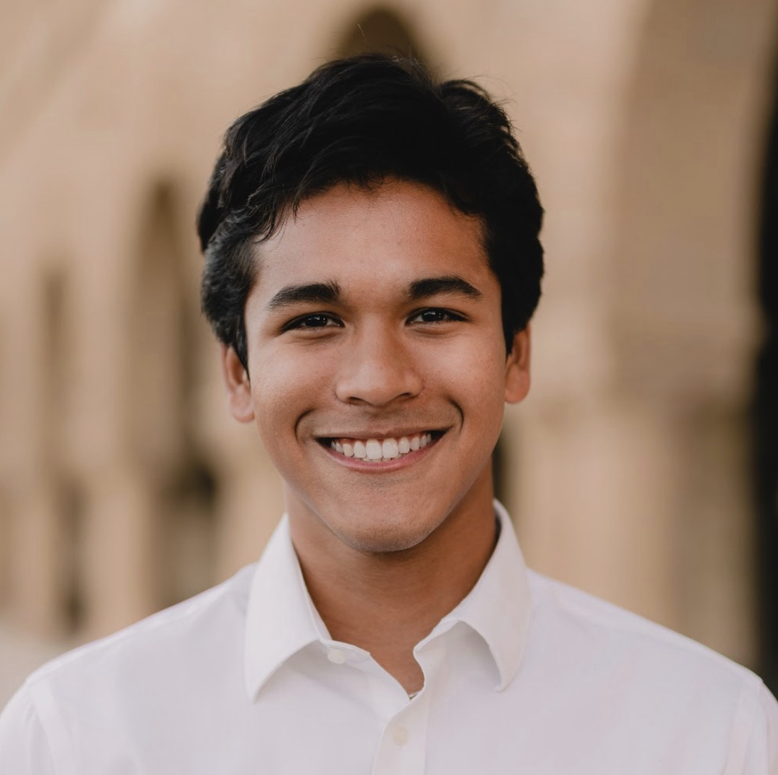 Young man with dark hair and a broad smile, wearing a white collared shirt, in an outdoor setting with blurred background.