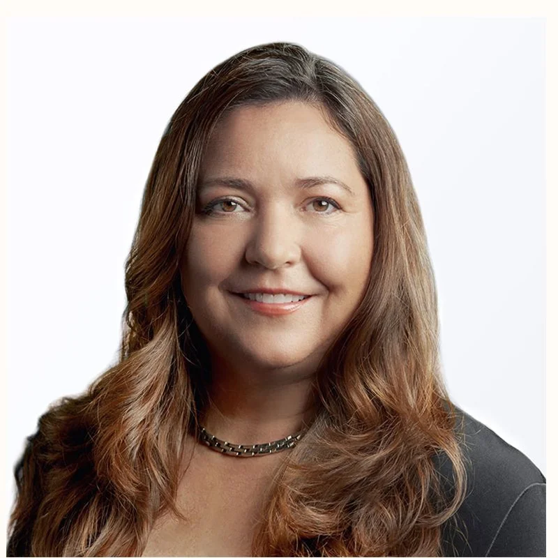 A woman with long brown hair smiling, wearing a black blazer and a chain necklace.