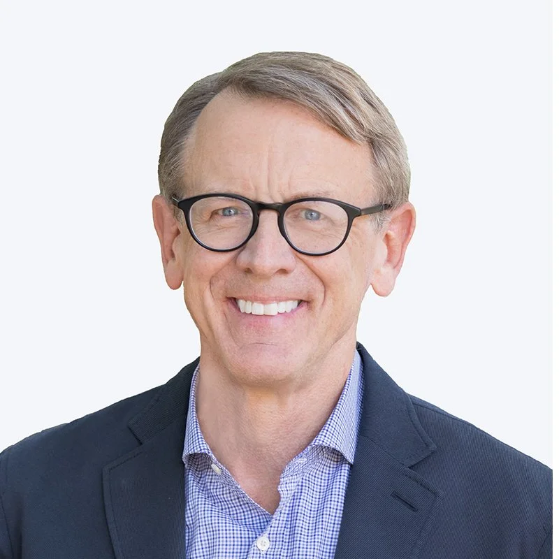 A middle-aged man with short light brown hair, wearing black glasses, a dark blazer, and a blue checkered shirt, smiling against a plain light background.