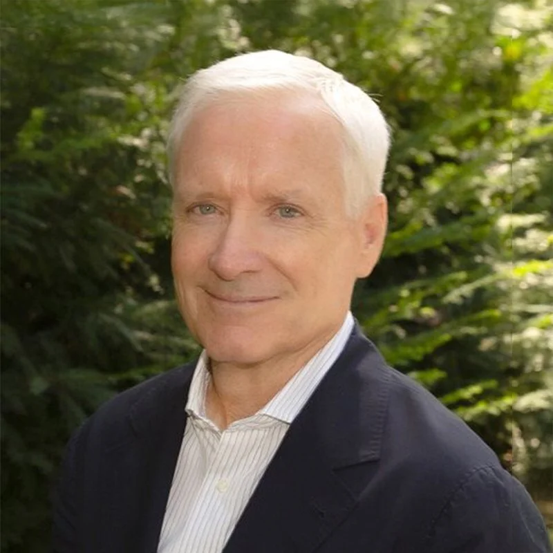 Portrait of an older man with white hair, wearing a dark blazer and a white collared shirt, standing outdoors with green foliage in the background.