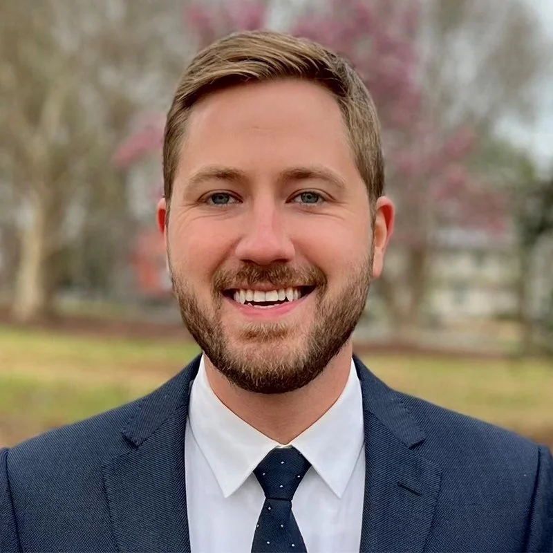 Headshot of a smiling man in a suit and tie outdoors with trees and a house blurred in the background.