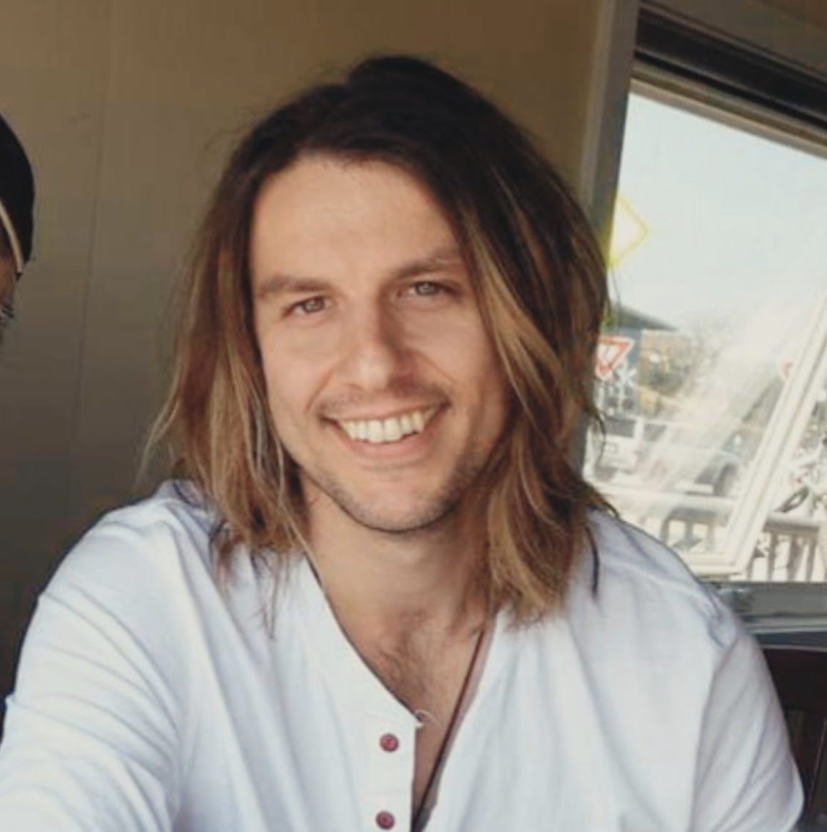 Smiling man with long brown hair, wearing a white shirt, sitting indoors near a window.