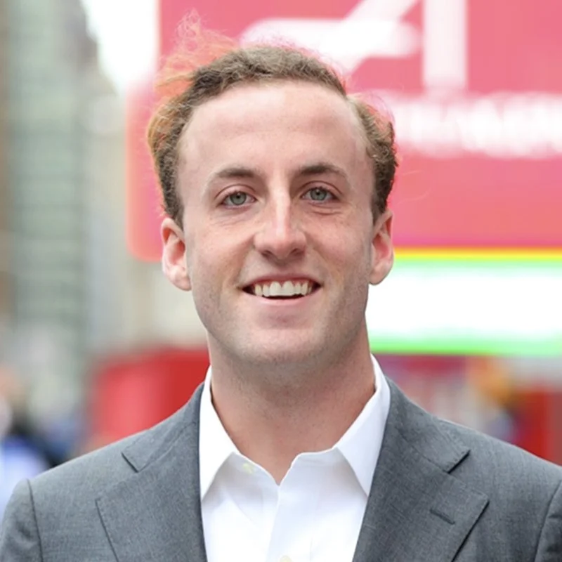 A young man with light skin, curly light brown hair, wearing a gray suit and white shirt, smiling outdoors with blurred city background and colorful signs.