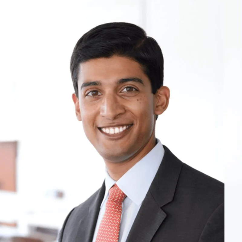 A smiling man wearing a dark suit, white shirt, and pink patterned tie in an office setting.