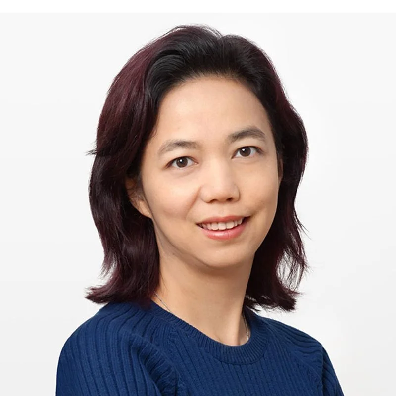 Portrait of a woman with shoulder-length dark red hair, wearing a blue sweater, smiling against a plain white background.
