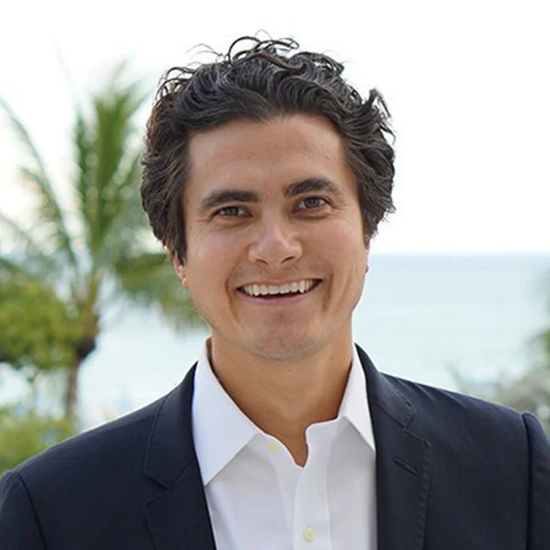 Smiling man in a suit with a background of palm trees and a body of water.