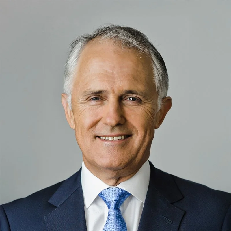 A smiling middle-aged man wearing a dark navy suit, white shirt, and light blue tie, standing against a plain gray background.