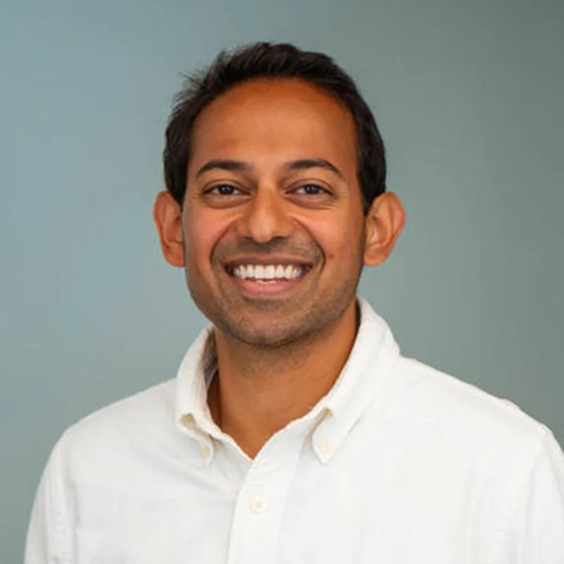 A smiling man with short dark hair wearing a white button-up shirt against a neutral background.