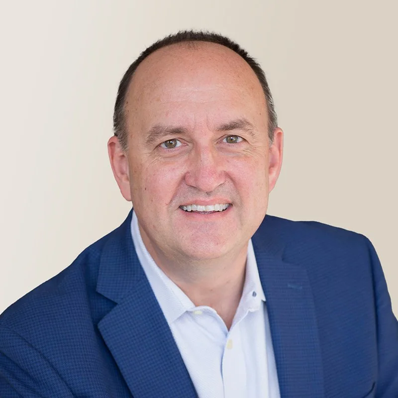 A smiling man with short dark hair and light skin, wearing a blue blazer and a white shirt, posing against a light background.