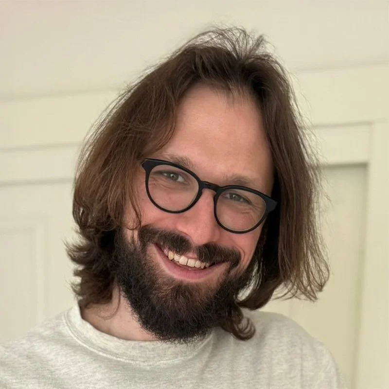 A smiling man with shoulder-length brown hair, glasses, and a beard, wearing a light-colored sweater, taking a selfie in front of a cream-colored wall.