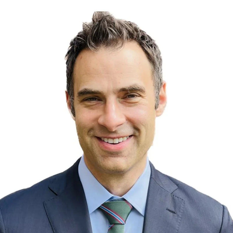 Professional headshot of a smiling man with short, dark hair, wearing a dark suit, light blue shirt, and striped tie against a white background.