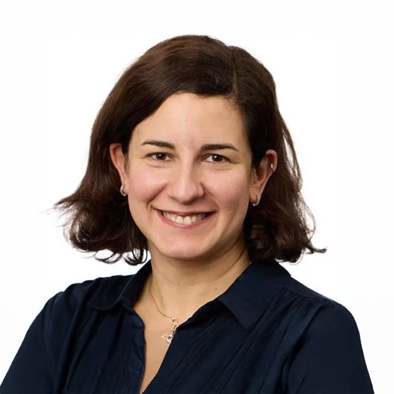 Headshot of a woman with shoulder-length brown hair smiling, wearing a dark navy blouse and jewelry, against a white background.